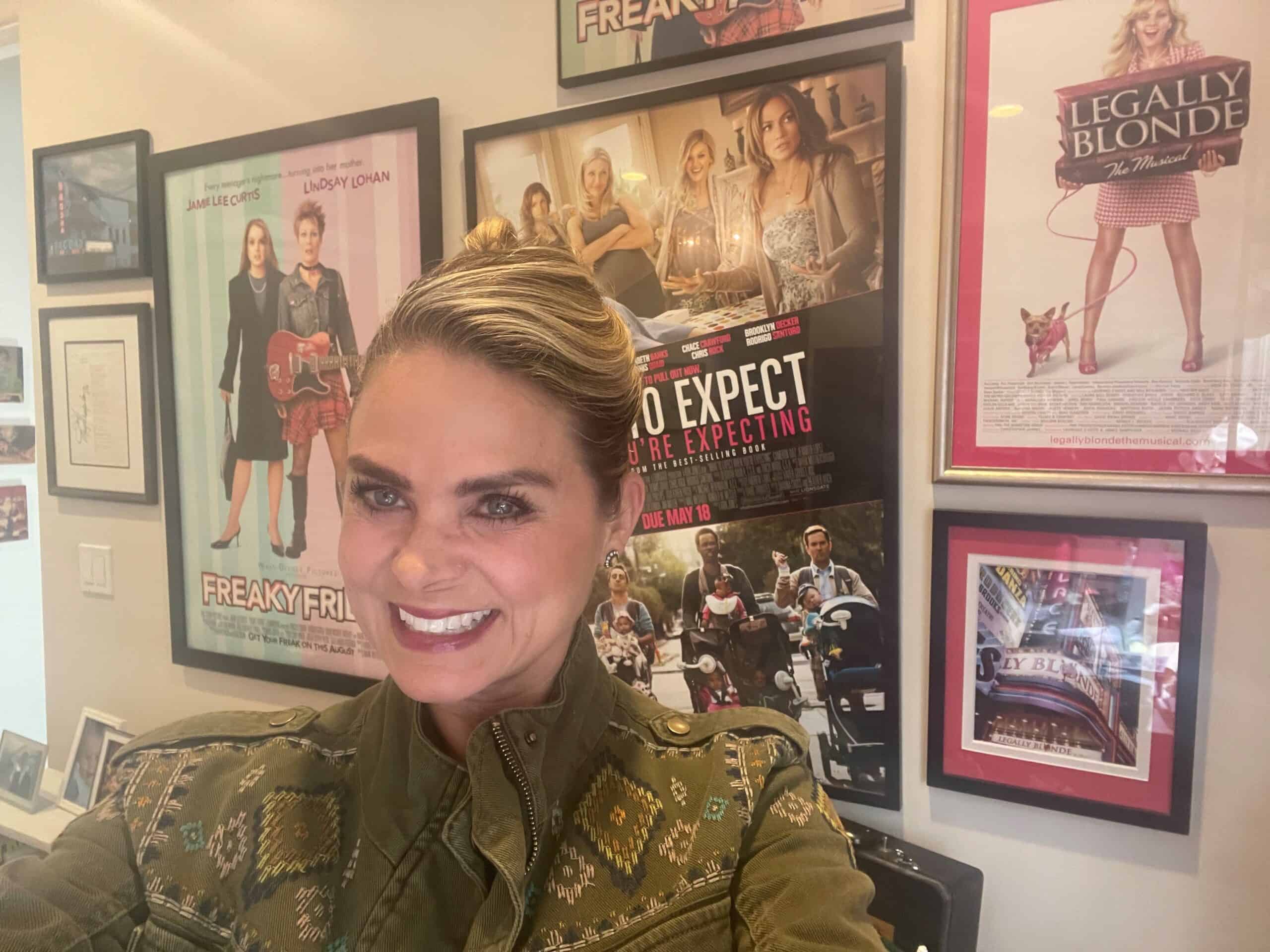 Heather Hach takes a selfie in front of a wall filled with framed movie posters, celebrating the art of cinema and embracing multifaceted female characters. Among the vibrant collection is a nod to Heather Hach's inspiring contributions to film.