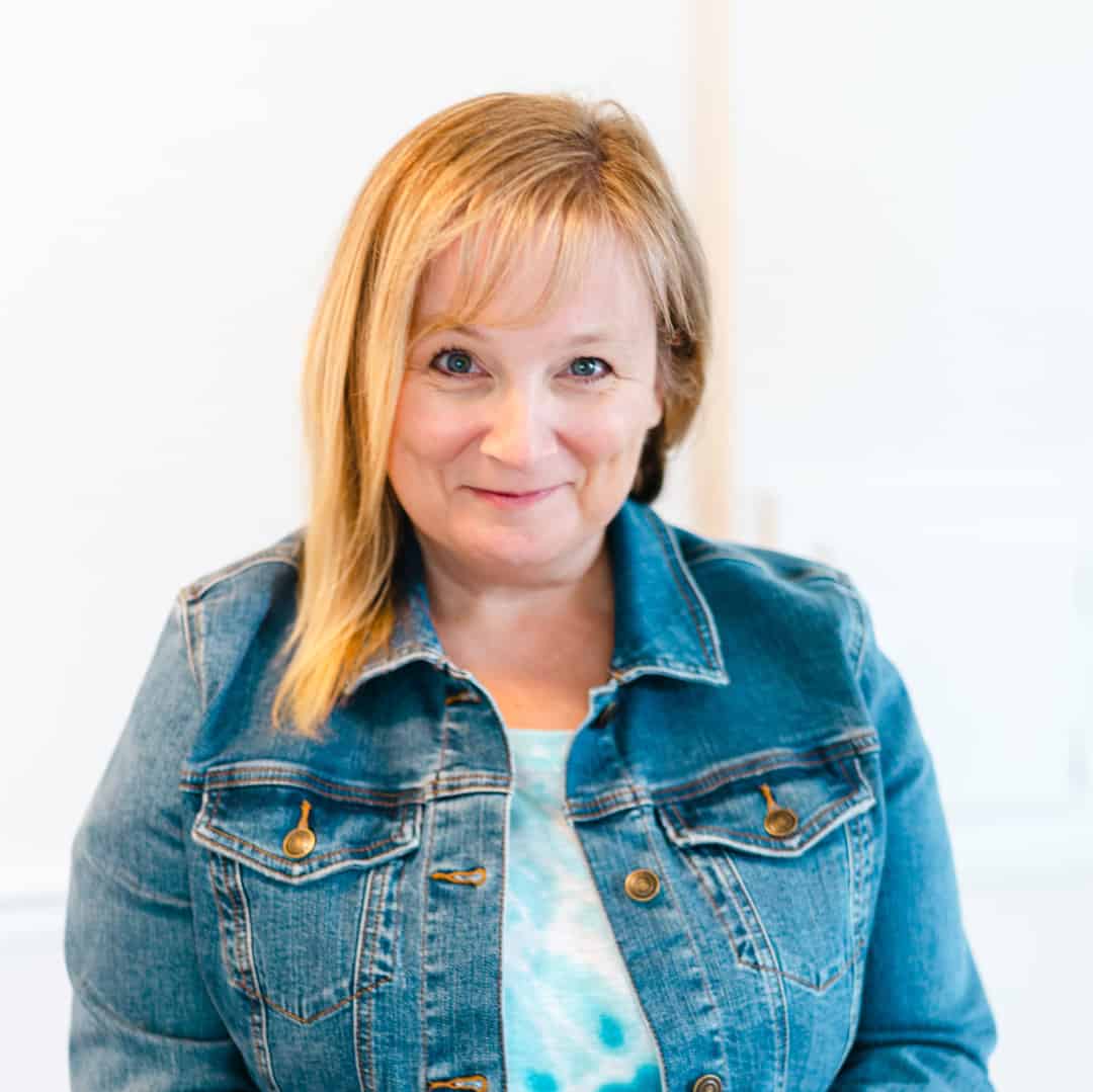 A person with blonde hair smiles, wearing a denim jacket over a blue shirt, set against a light background. They radiate hope and resilience, echoing themes from "Breaking Cycles of Abuse with Kathy Wisniewski.