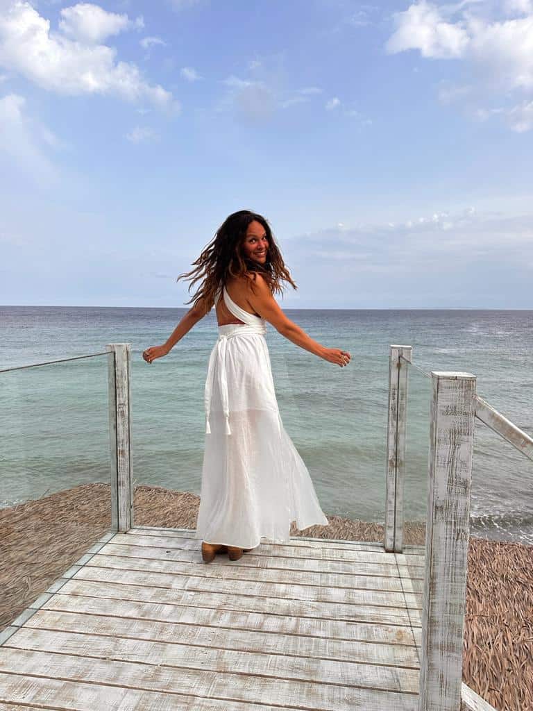 A woman in a white dress stands on a wooden deck by the sea, joyfully embracing the healing power of movement with Cortney McDermott. She smiles under the partly cloudy sky, embodying graceful serenity.