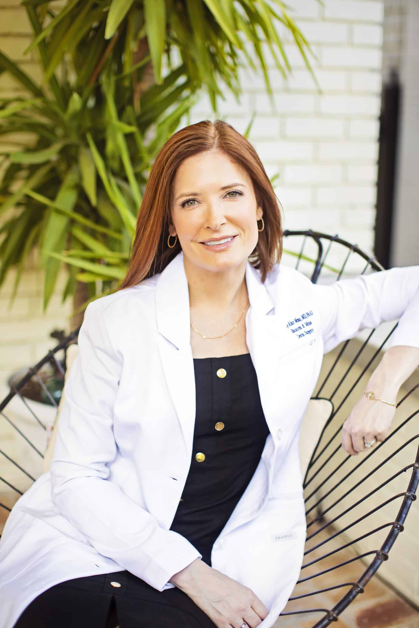 Dr. Mary Alice Mina in a white lab coat, specializing in menopause skin changes, sitting on a metal bench and smiling at the camera.