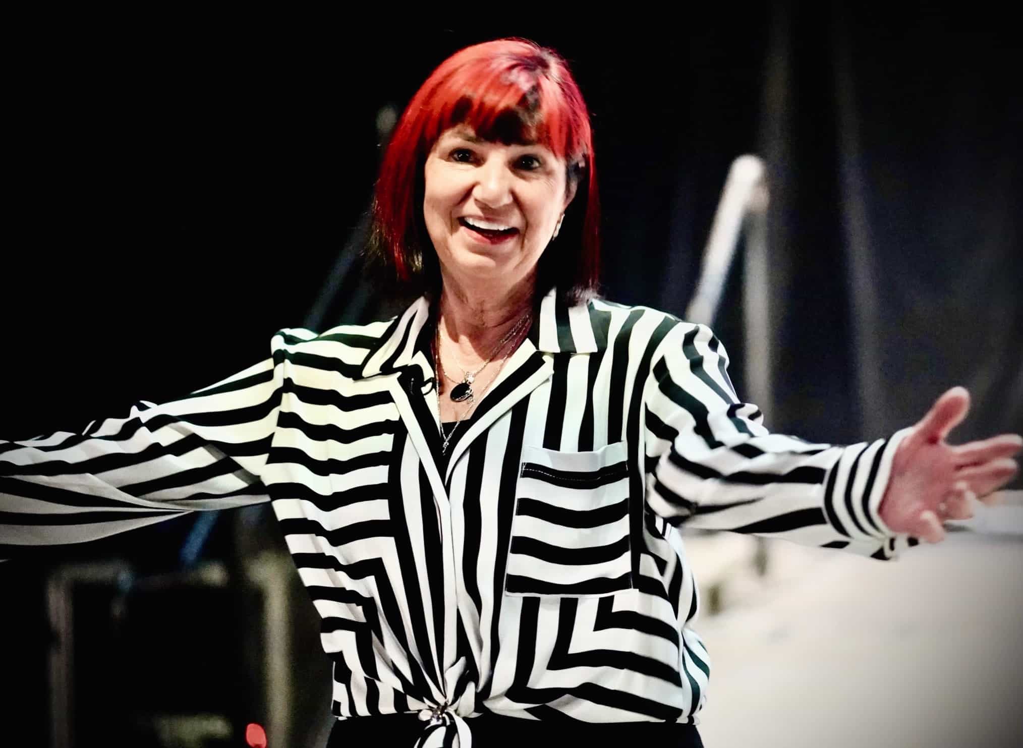 Gail Taylor, wearing a black and white striped blouse, is smiling and extending her arms as if to welcome someone or gesture during a presentation on her midlife pivot: from finance to music