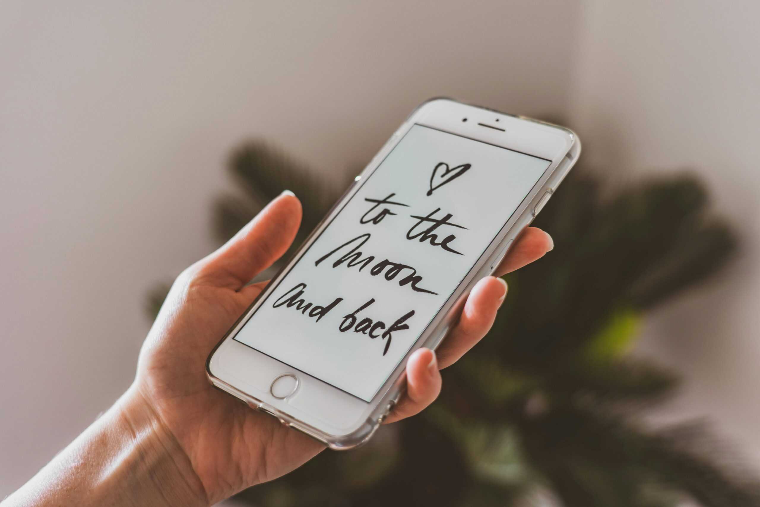 A hand holding a smartphone displaying the message "to the moon and back" with a heart and blurred plant background, symbolizing effective communications.