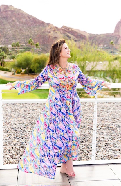 Elaine Glass in a colorful, patterned dress stands barefoot on a balcony with mountains in the background, looking away as she spreads her arms, embodying her pivot to purpose.