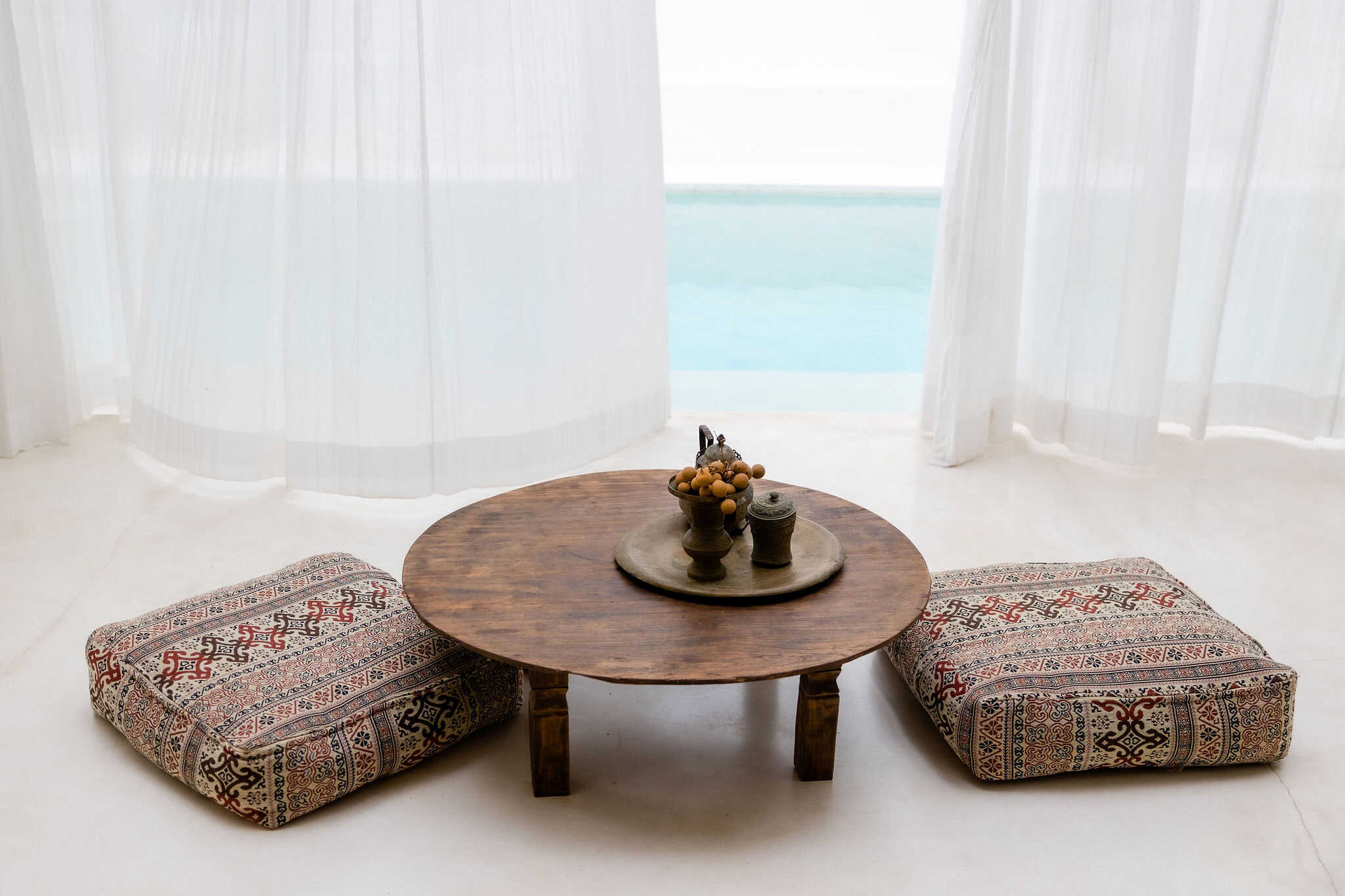A cozy seaside relaxation spot embracing a holistic path, with a wooden table, patterned floor cushions, and sheer curtains overlooking a tranquil ocean view.