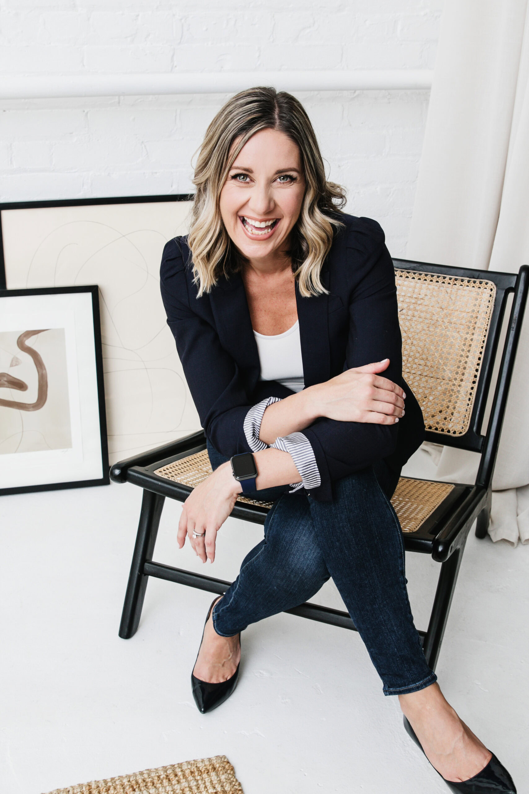 A smiling woman named Shannon Talbot in a navy blazer and striped shirt sits on a black chair in a white room, with framed artwork leaning against a wall nearby.
