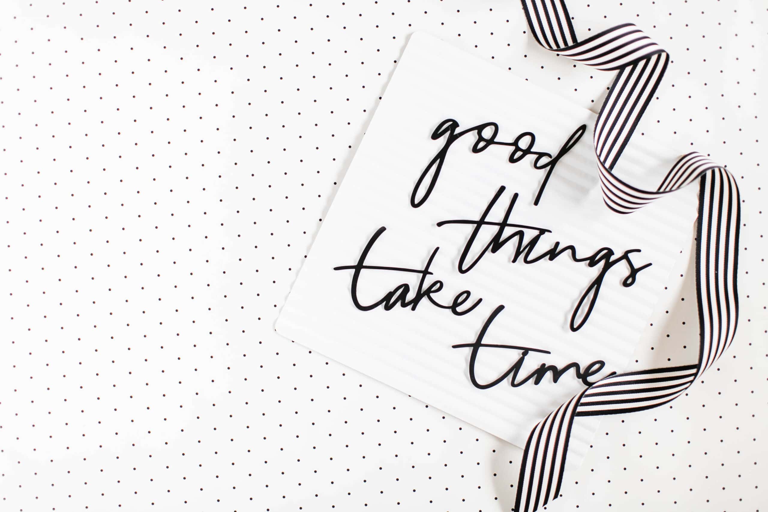 A white board with the inspirational quote "good things take time" from stories of empowerment, written on it, placed on a dotted background with a striped ribbon.
