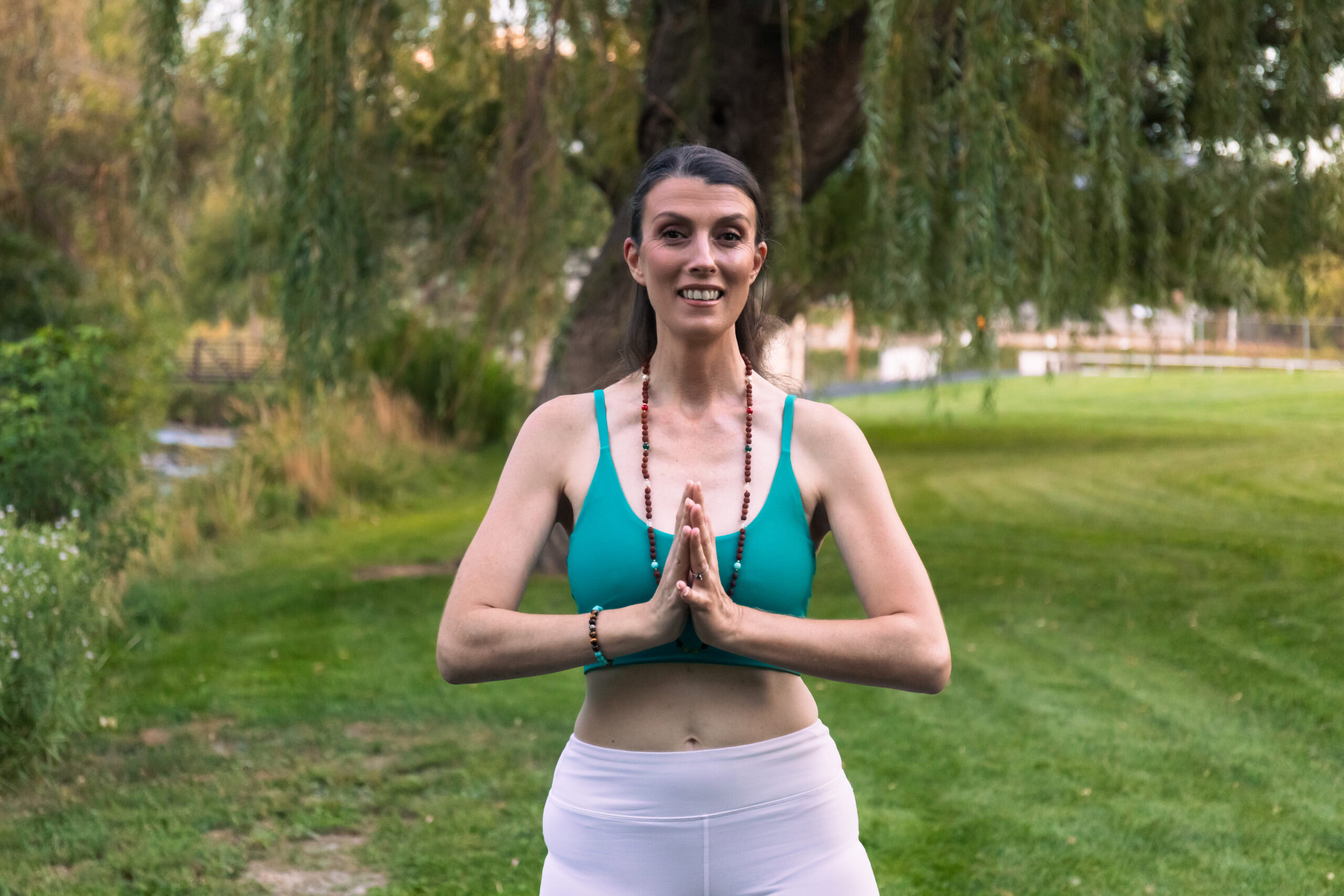 Danae wearing a green top and white leggings stands outside in a yoga pose with hands pressed together, practicing Danae Robinett Breathwork against a backdrop of greenery and trees.