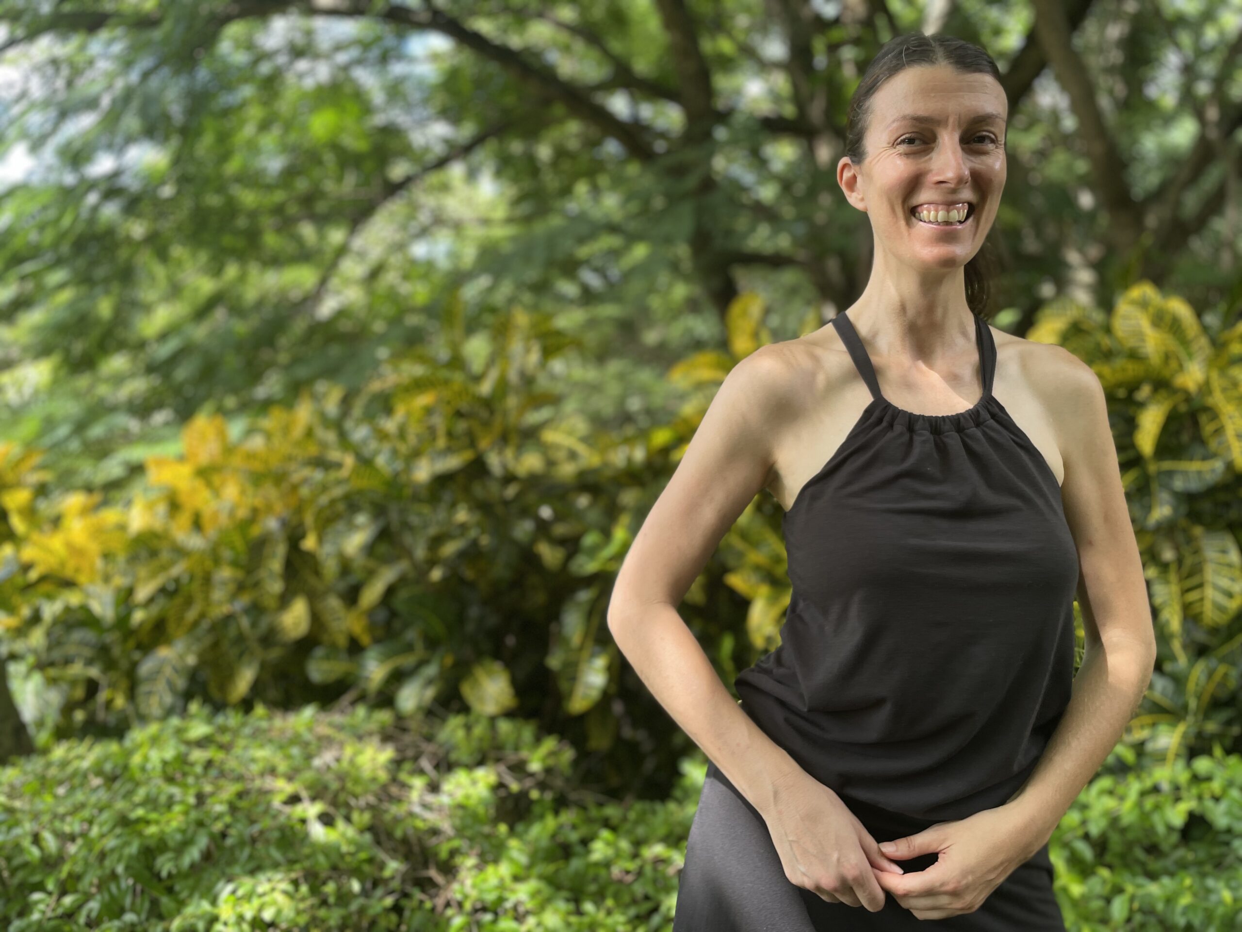 Danae Robinett is standing outdoors, smiling with green foliage and yellow leaves in the background, wearing a sleeveless black top.