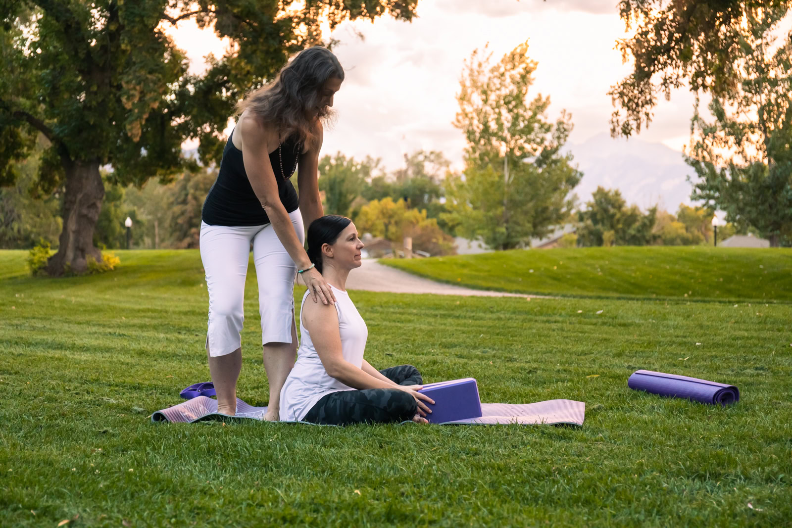 Two people are practicing yoga in an outdoor park; one is sitting on a mat with a yoga block, while Danae Robinett stands behind, assisting with a shoulder adjustment. Danae Robinett's techniques are empowering women through breath and mindful movements.