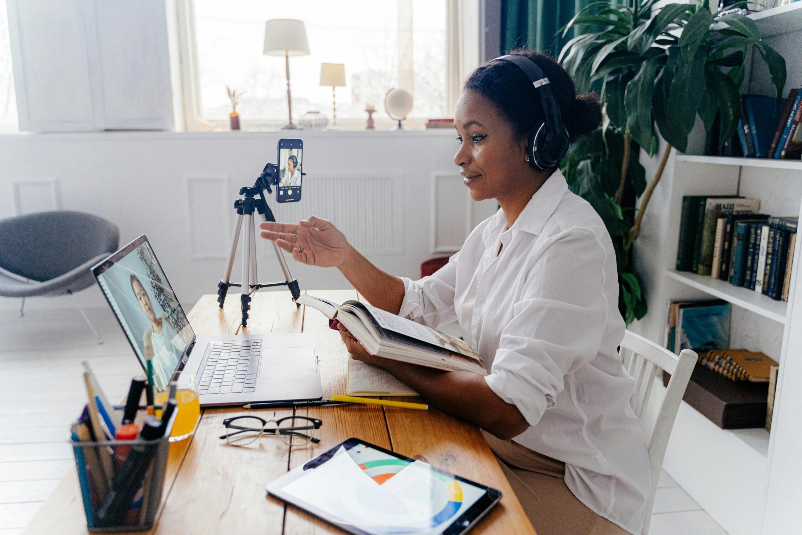 A woman embracing gadgets in midlife uses headphones and a smartphone on a tripod to film herself for a video call while consulting a book and interacting with a laptop.