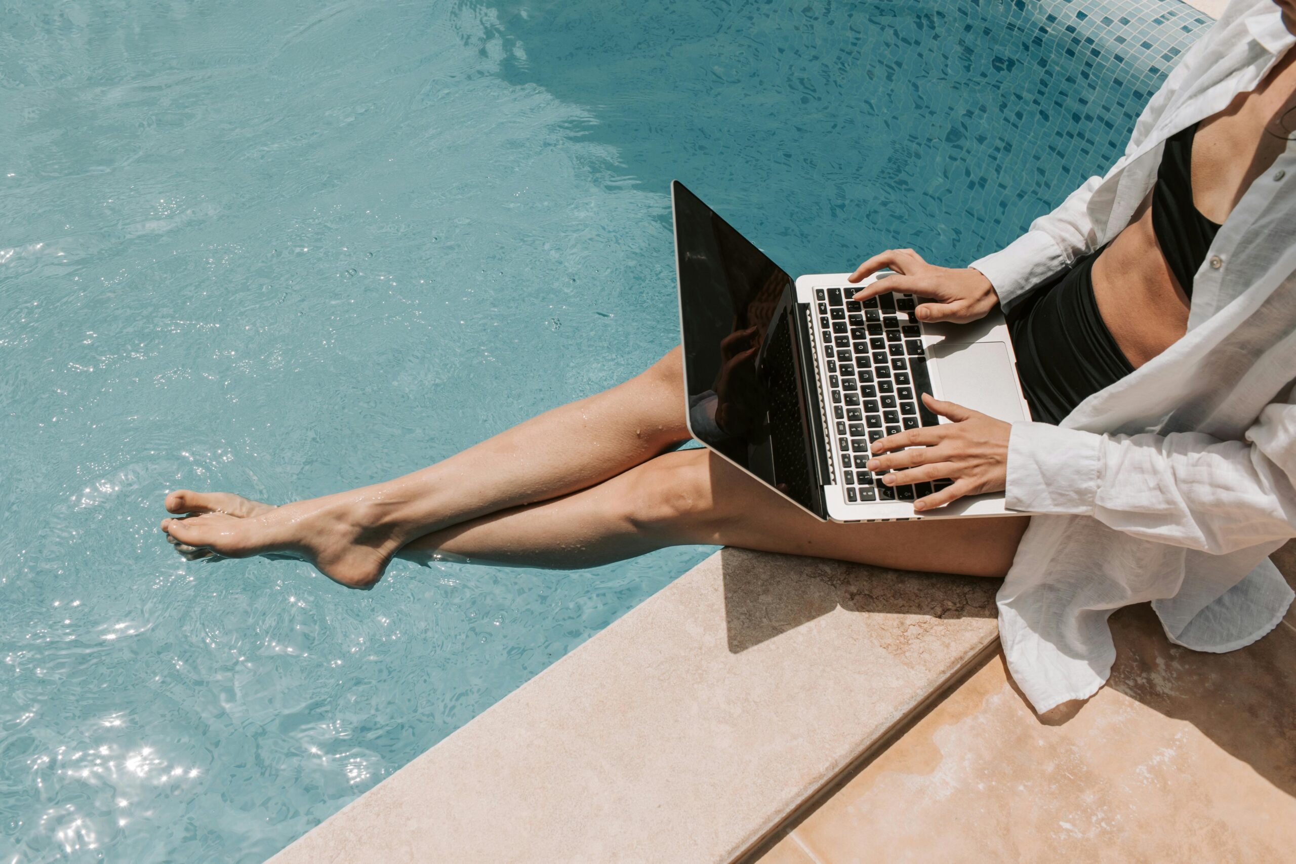 A person practicing smart living by using a laptop by a pool's edge, with legs dipped in water, dressed in a white shirt and dark shorts.