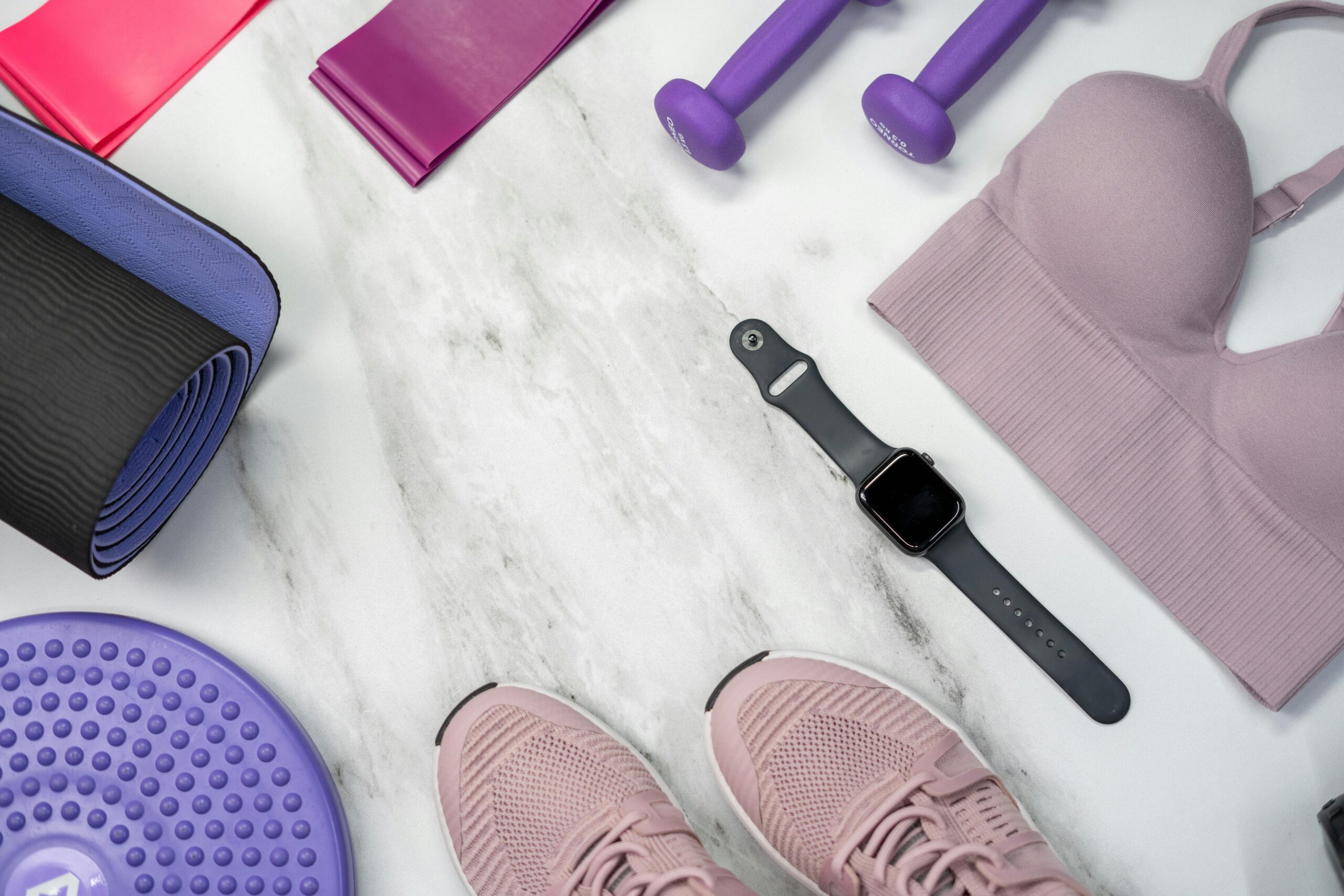 Top view of workout equipment on a marble floor, featuring pink sneakers, a yoga mat, dumbbells, a sports bra, a smartwatch, and other health accessories.