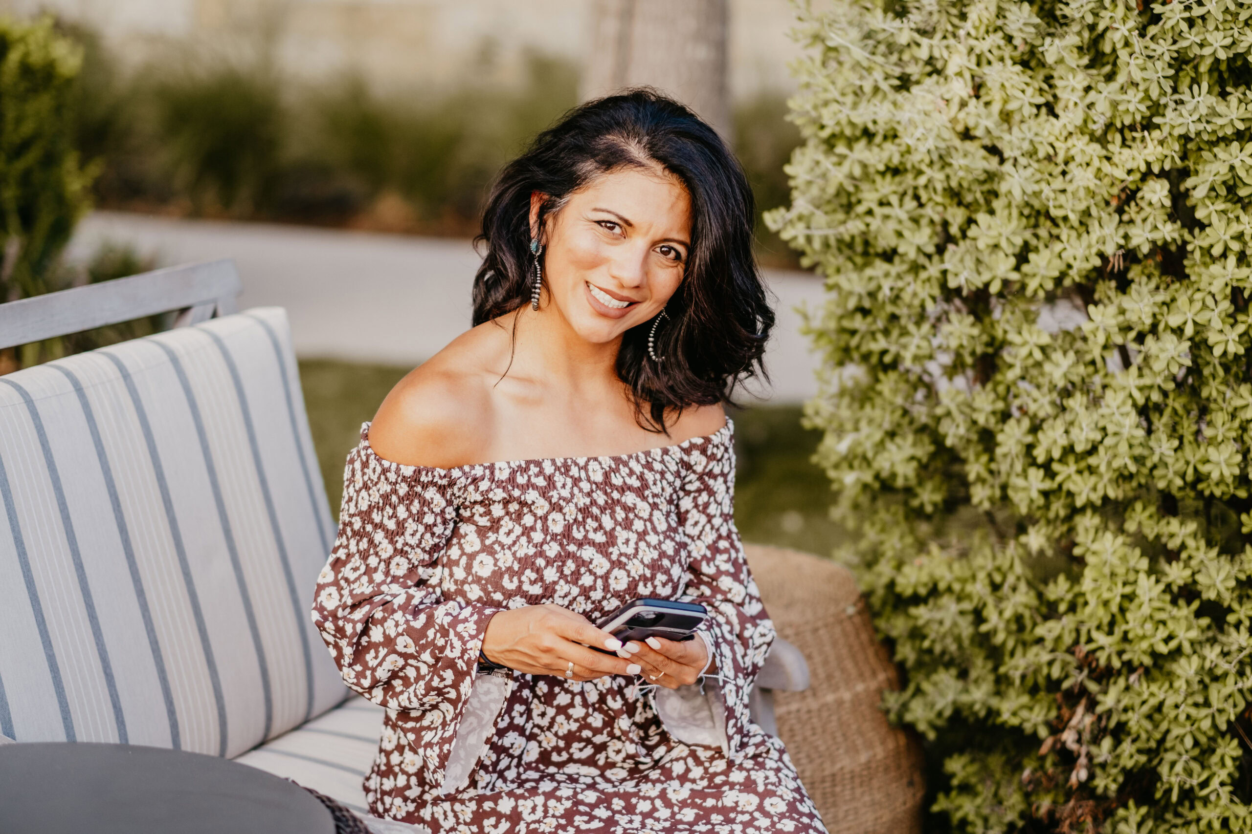 Susie Maldonado sits on an outdoor bench, wearing a floral off-the-shoulder dress, holding a phone, and smiling at the camera. She is surrounded by greenery, embodying the essence of thriving in midlife.