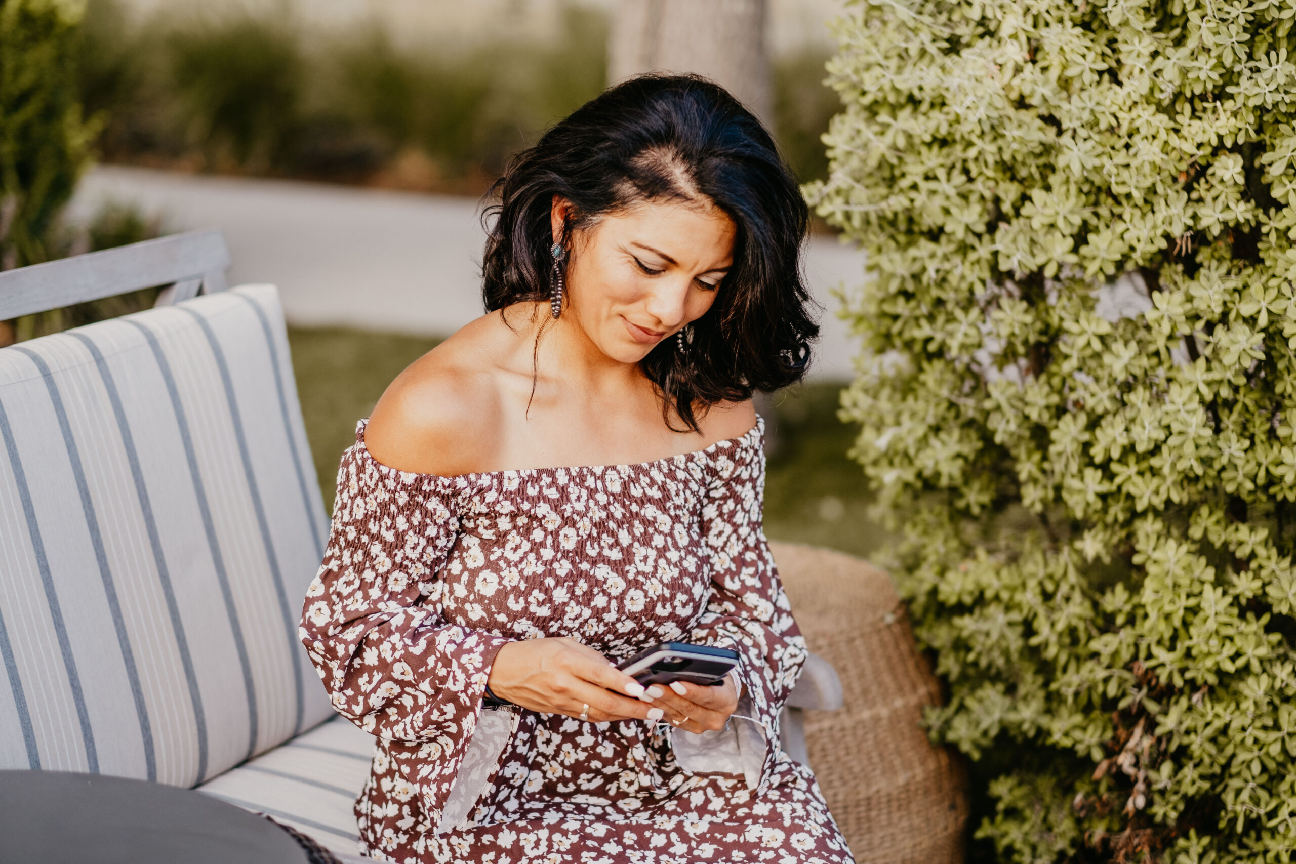 Susie Maldonado with shoulder-length dark hair sits on an outdoor bench, looking at her smartphone. She wears an off-shoulder, floral dress, with green foliage in the background. Amidst thriving greenery, she seems deep in thought, perhaps pondering the transformative secrets of midlife.