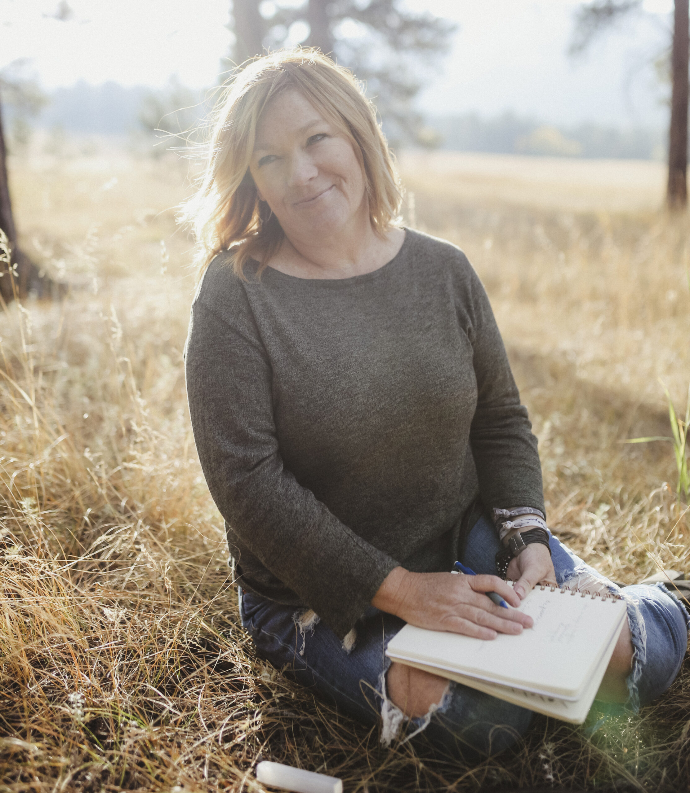 Wendy Wright sitting in a field journaling in a notebook with a happy expression, surrounded by tall grass and trees, with sunlight filtering through.