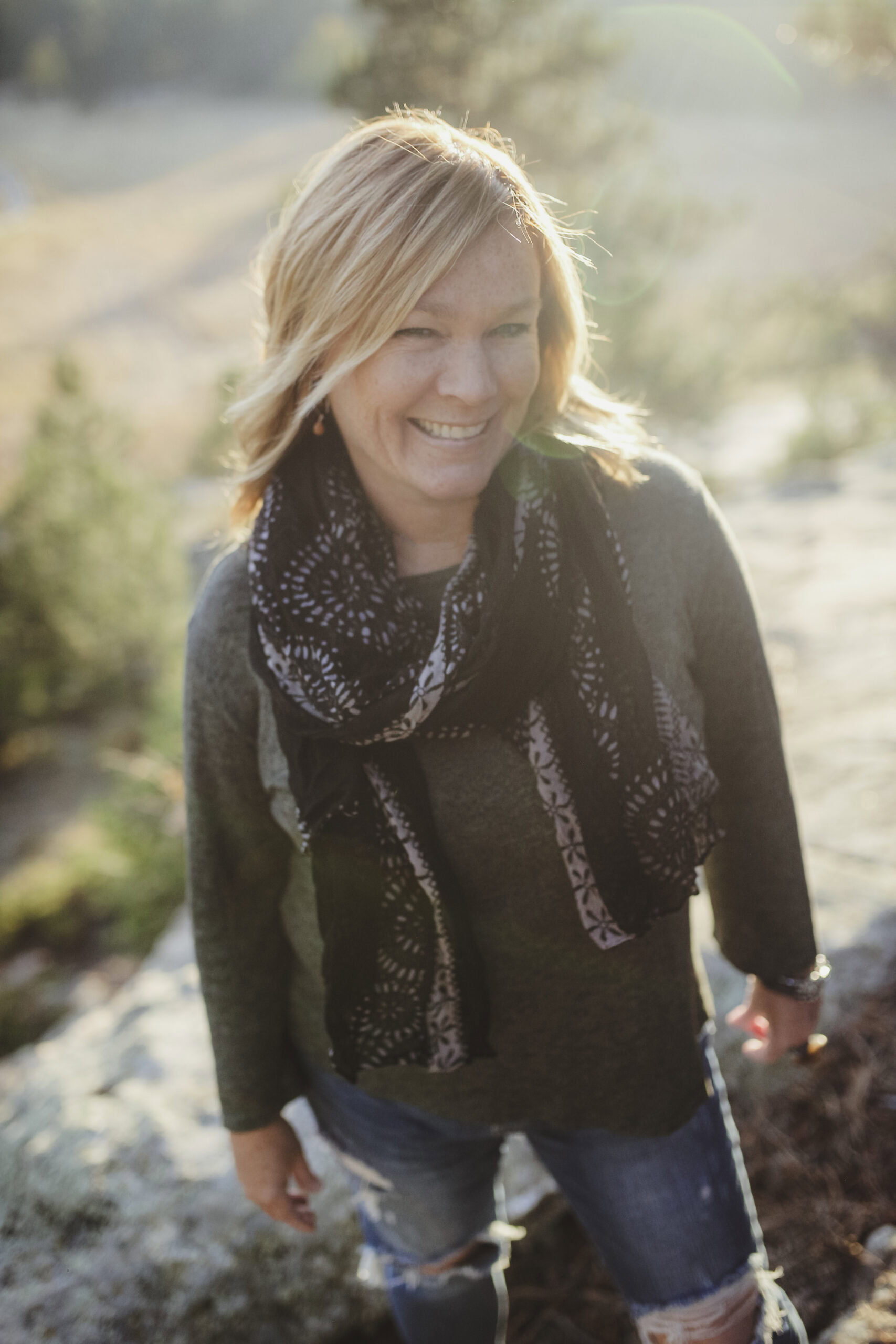 A Wendy Wright smiling outdoors, wearing a scarf, ripped jeans, and a sweater, with sunlight and a financial therapy book in her hand against the background of a natural landscape.