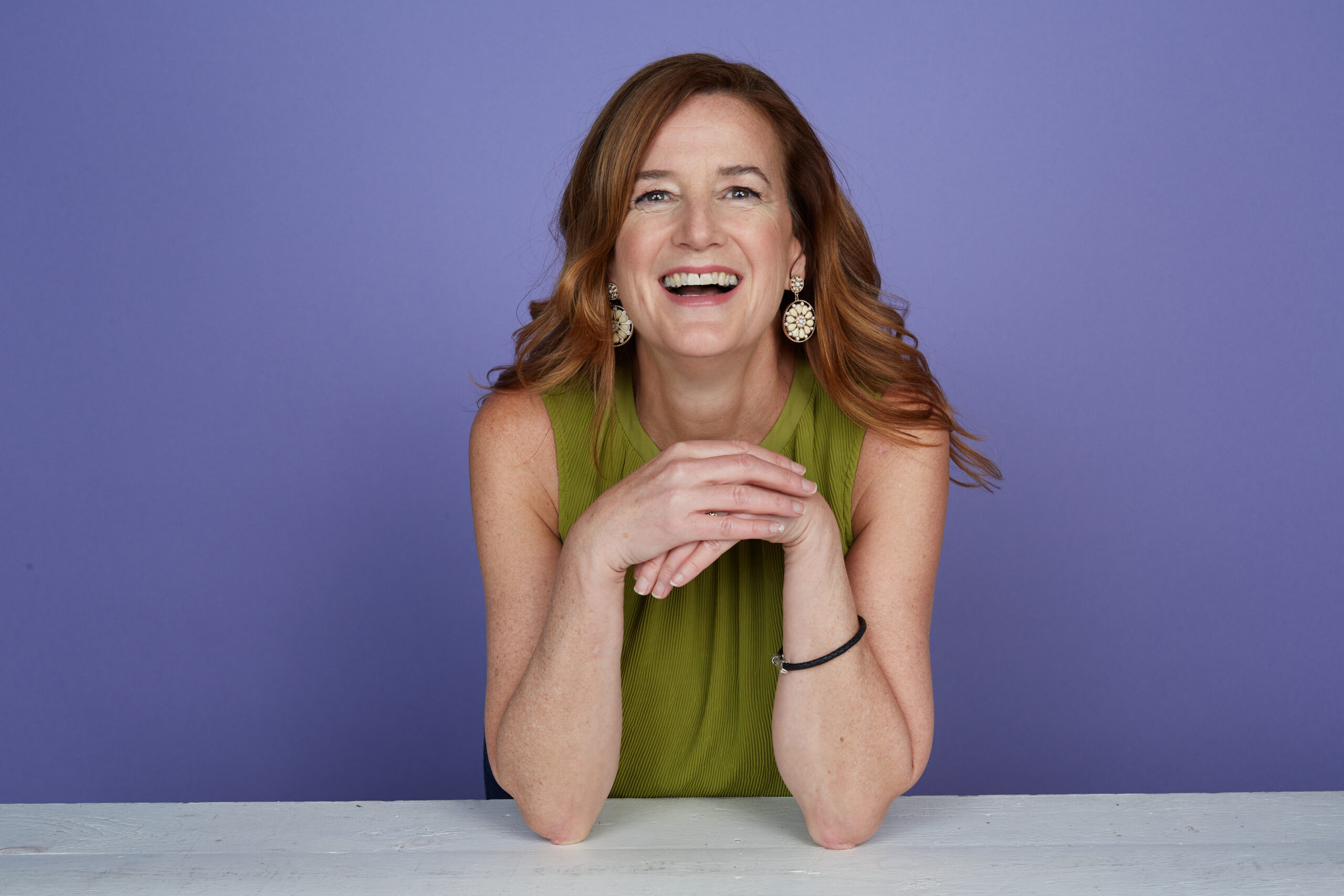 Annie Gaudreault, a woman with long red hair, wearing a green sleeveless top, smiles while leaning on a white table against a purple background.