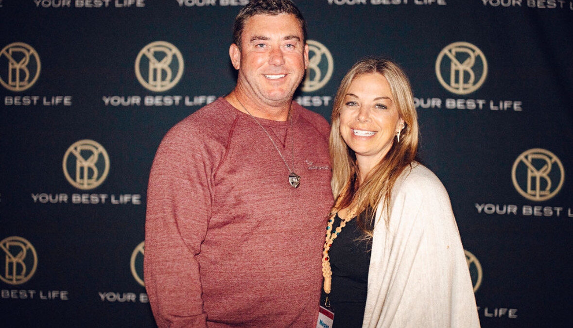 A man and a woman are standing together, smiling, in front of a backdrop that reads "YOUR BEST LIFE" with a circular logo. The man is wearing a red shirt, and the woman, Megan McShane, is wearing a white shawl. They embody the spirit of thriving in midlife and reigniting your passions.