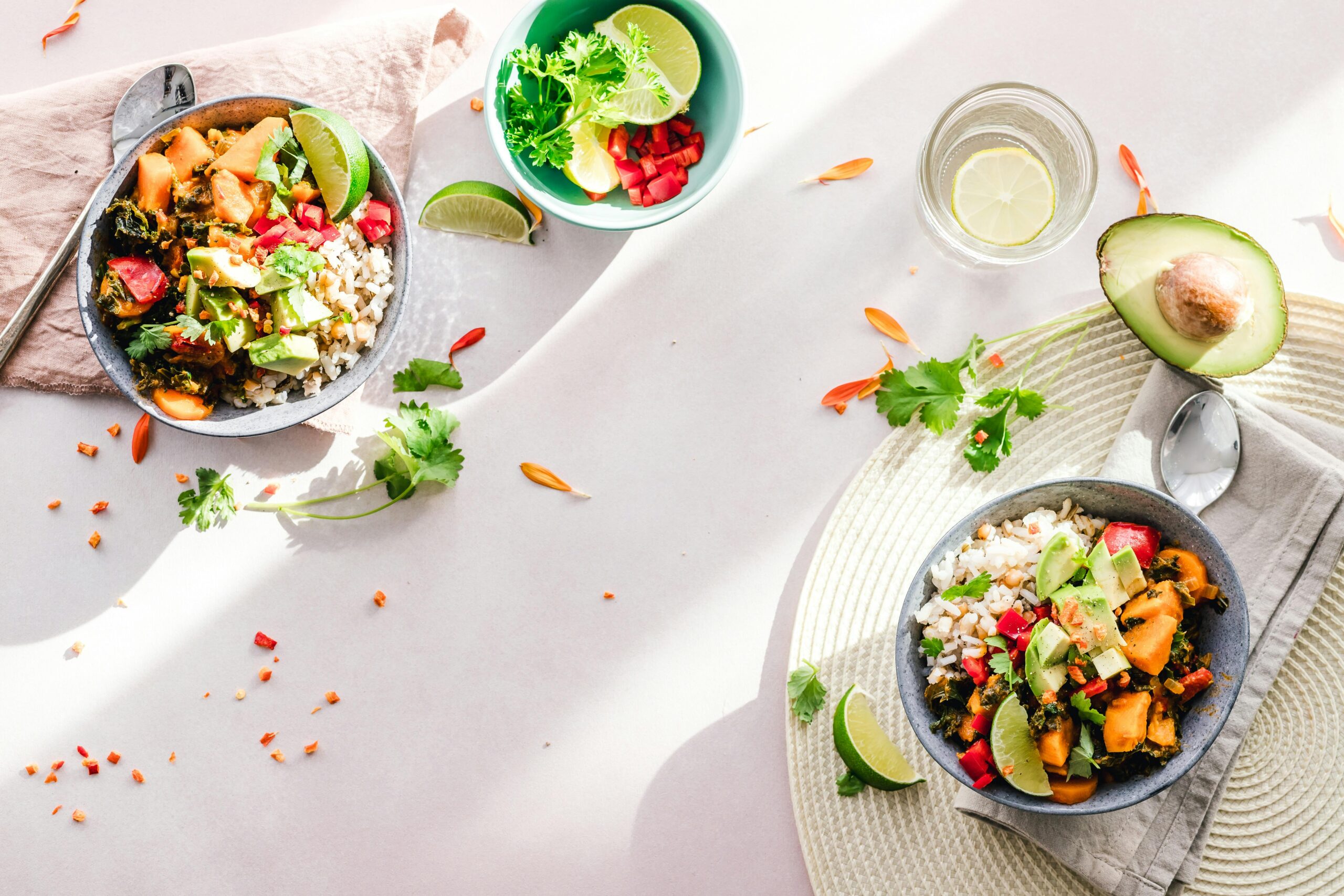 Two bowls of colorful food including avocado, rice, and vegetables are on a light surface with scattered herbs and lime wedges. Nearby, a bowl of lime slices and a glass of lemon water reflect the vibrancy of community dining.