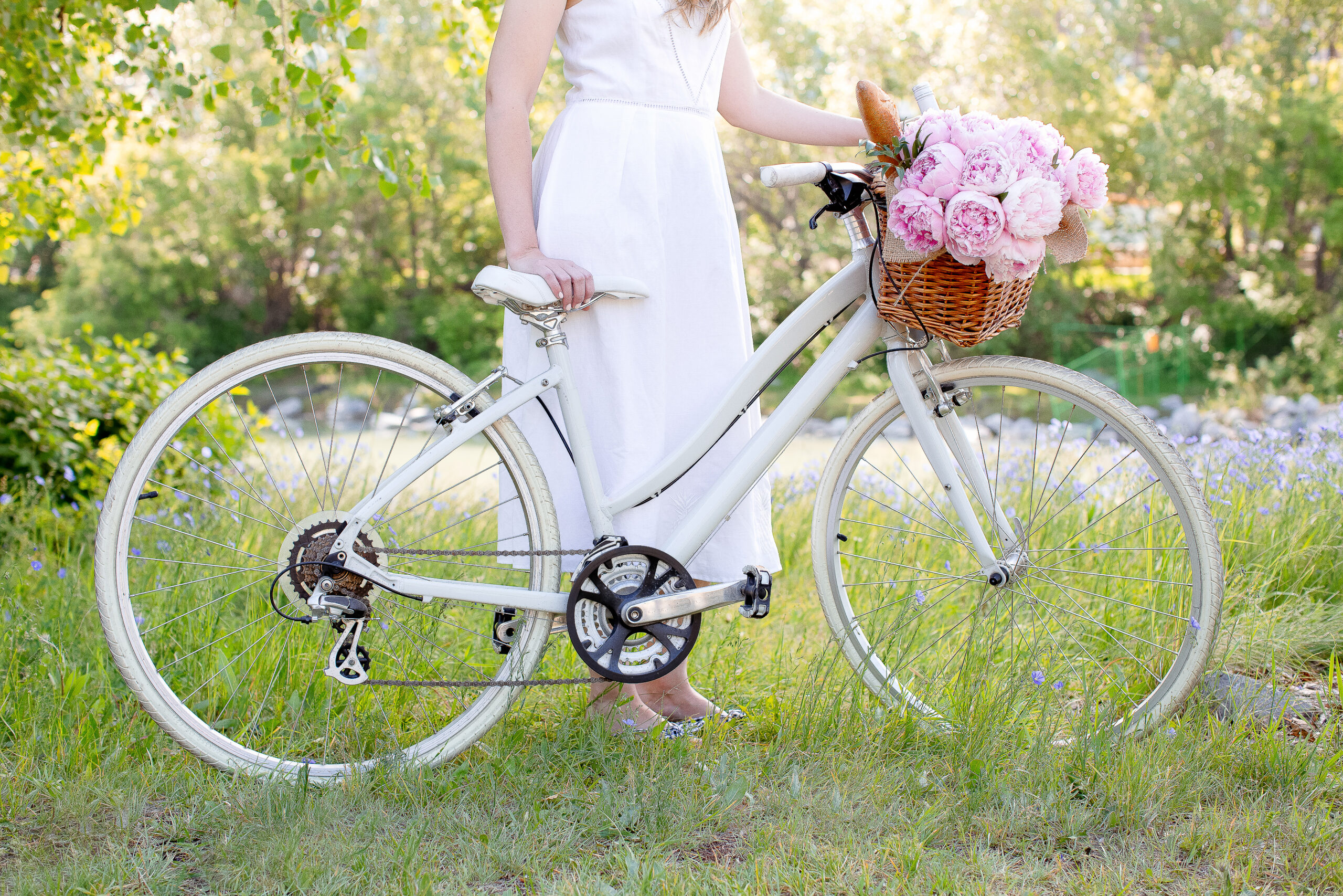 A person in a white dress stands beside a white bicycle with a basket full of pink flowers and baguettes, embracing the great outdoors in a grassy area.