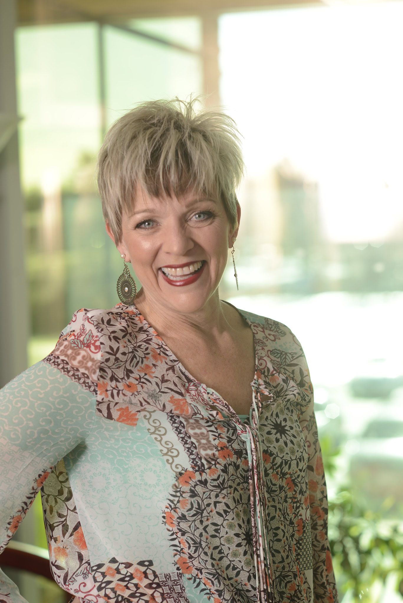 Dr. Kris smiling person with short blonde hair, wearing a patterned blouse and earrings, stands in a bright room with large windows, embracing functional medicine.