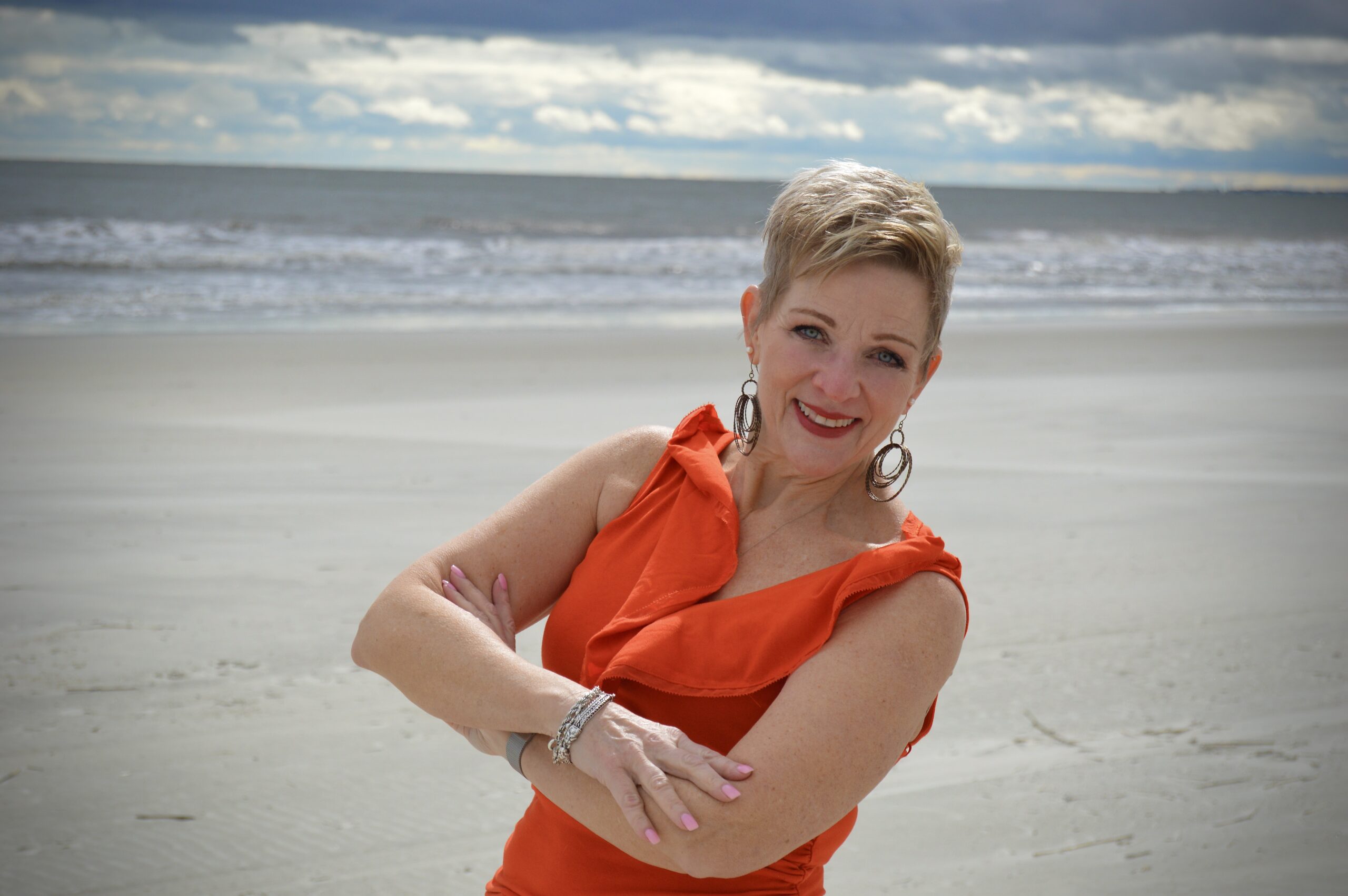 Dr. Kris standing on a sandy beach, posing with folded arms. They are wearing an orange sleeveless top and large hoop earrings. The sea and cloudy sky in the background create a serene setting, highlighting the impact of lifestyle choices.