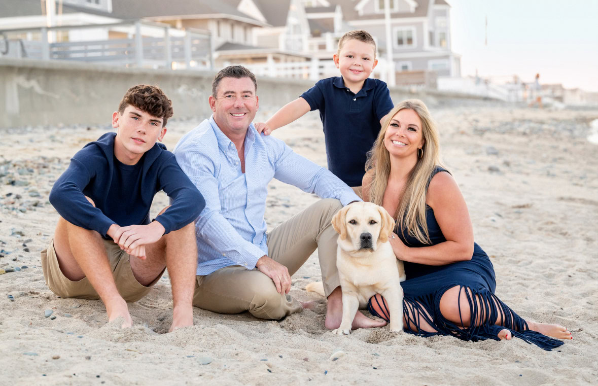 Megan McShane's family of four and their dog sit together on a sandy beach with houses in the background, engaging in community and connection.