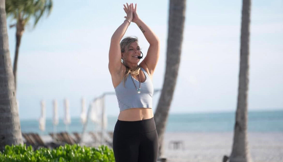 Megan McShane, in athletic wear, stands on a beach with arms raised, wearing a headset microphone. Palm trees and the ocean form the background as she shares her secrets to reigniting your passions and thriving in midlife.