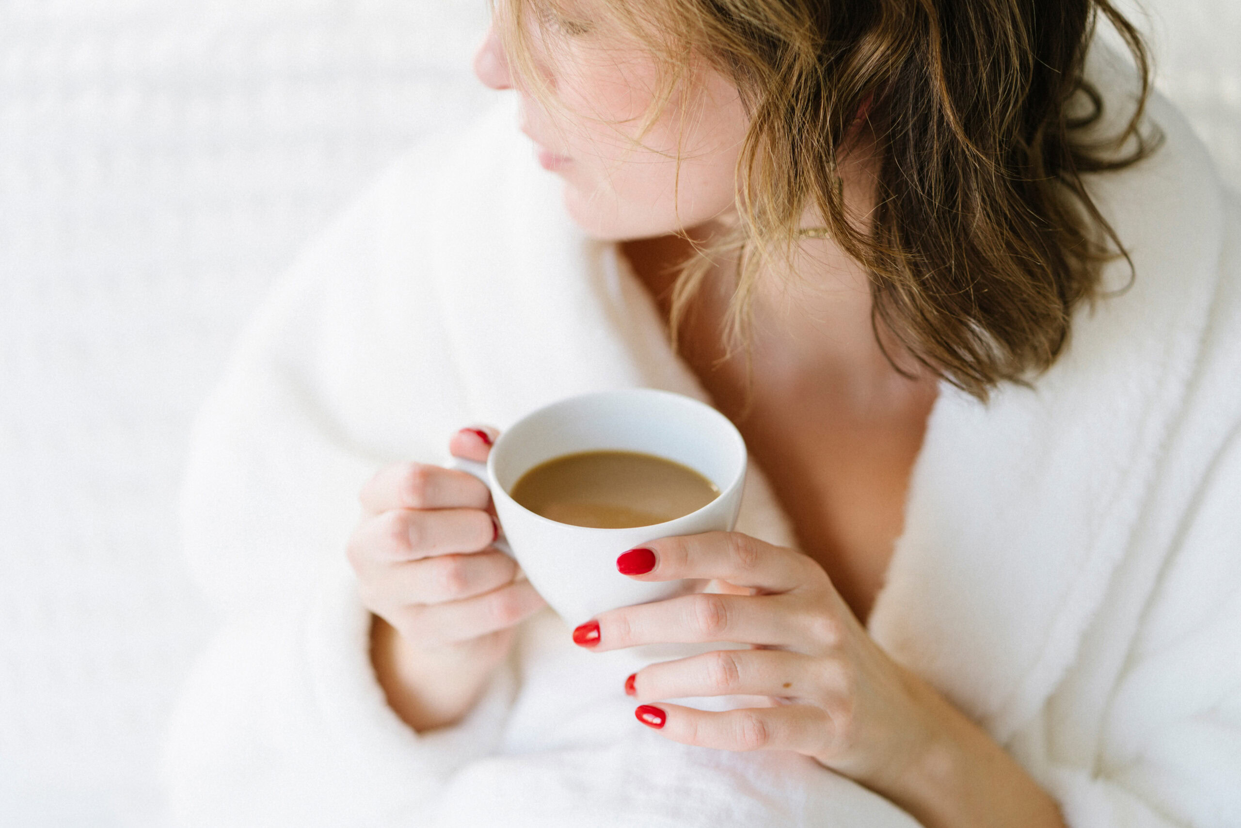 A person with red nail polish holds a white cup of coffee, wearing a white robe, and gazes into the midlife landscape, looking away from the camera.