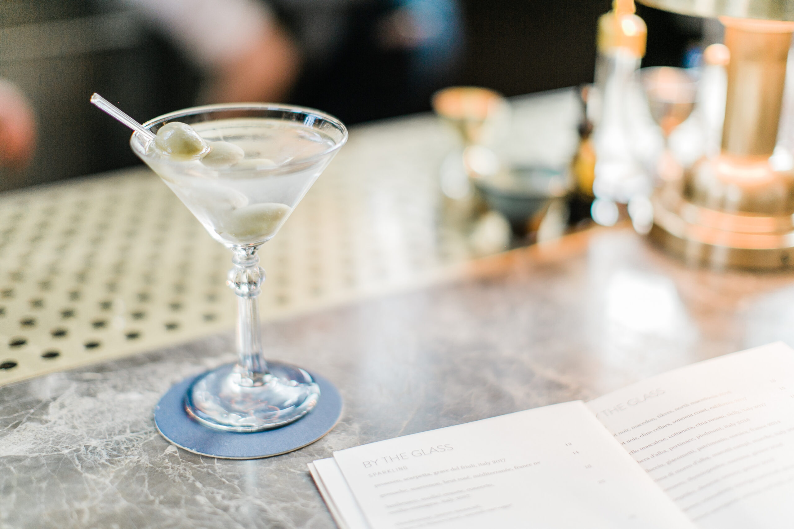 A martini with olives on a stirrer sits on a bar counter next to an open menu, offering the perfect moment for some quiet journaling.