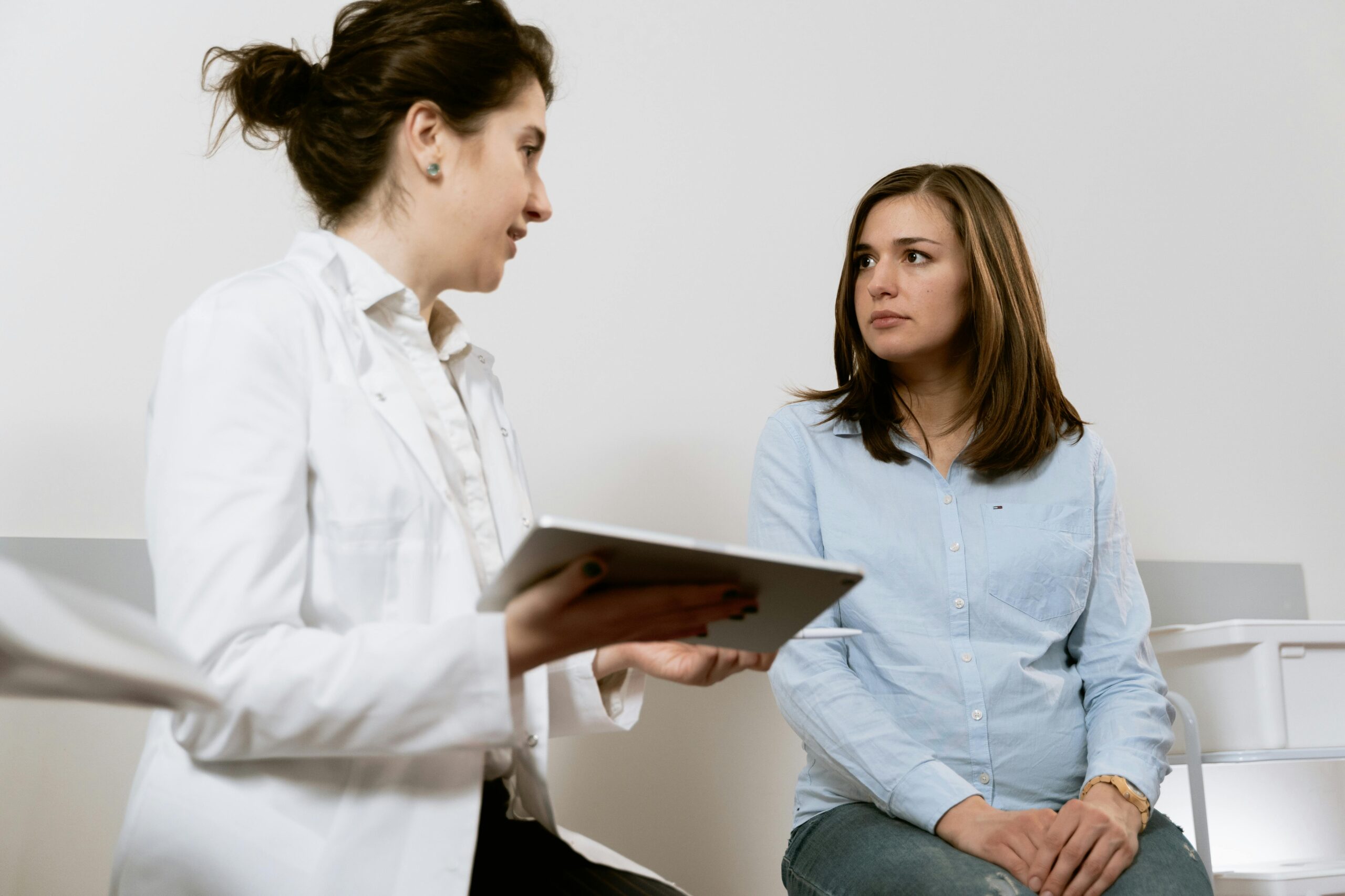 A healthcare professional holding a tablet converses with a seated woman in casual clothing, partnering with health care providers to ensure comprehensive care in a clinical setting.