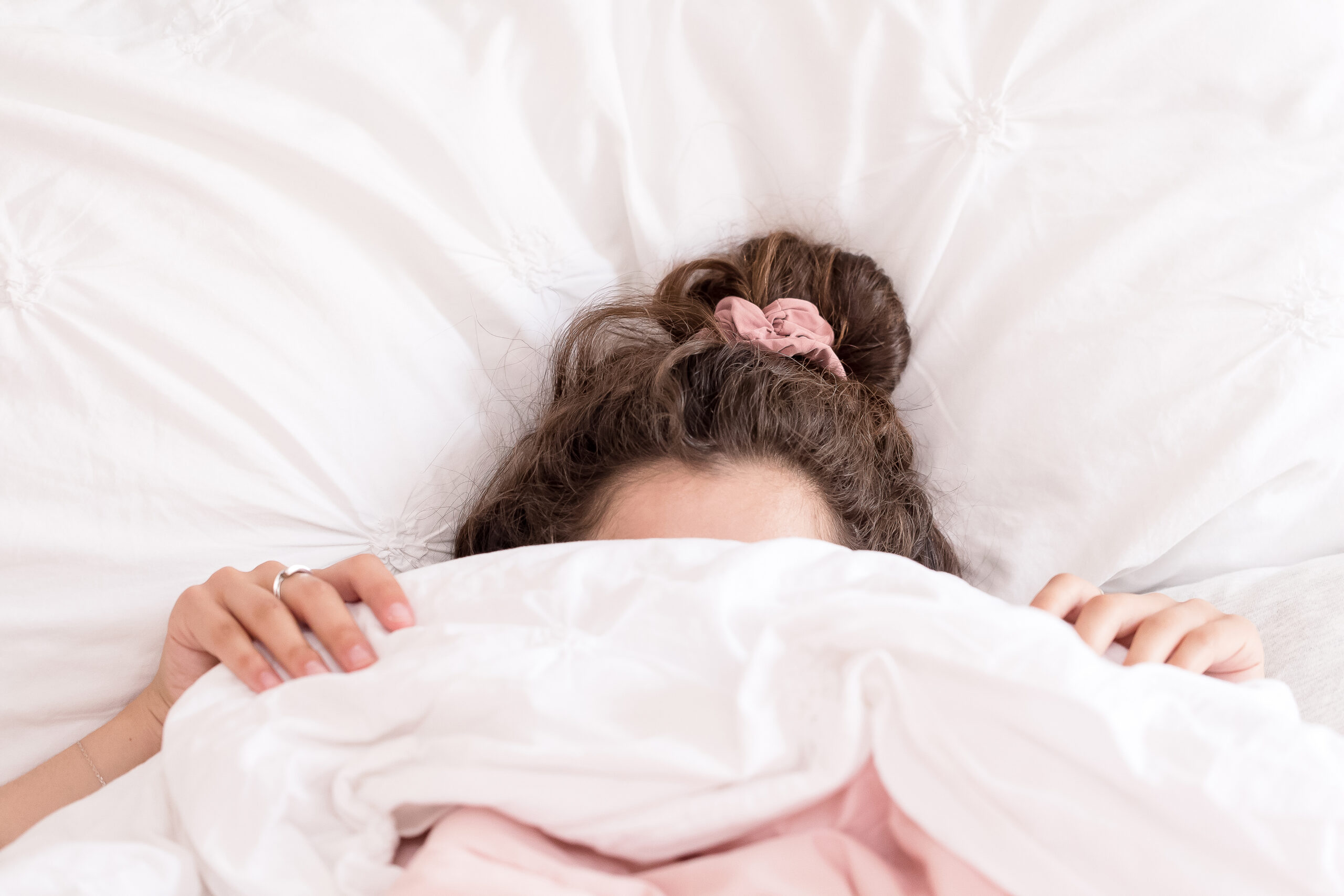 A person lies in bed partially hidden under white blankets with only the top of their head and hands visible. They have a pink scrunchie in their hair, perhaps reflecting a moment of relaxation after meditation.