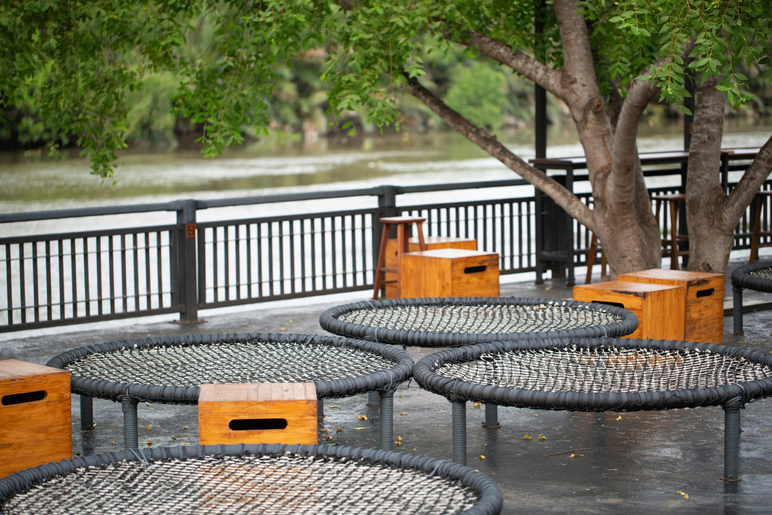 Outdoor seating area with round rope chairs and wooden side tables, situated near a metal railing along a riverbank. Experience the joy of movement while trees with green foliage provide shade.