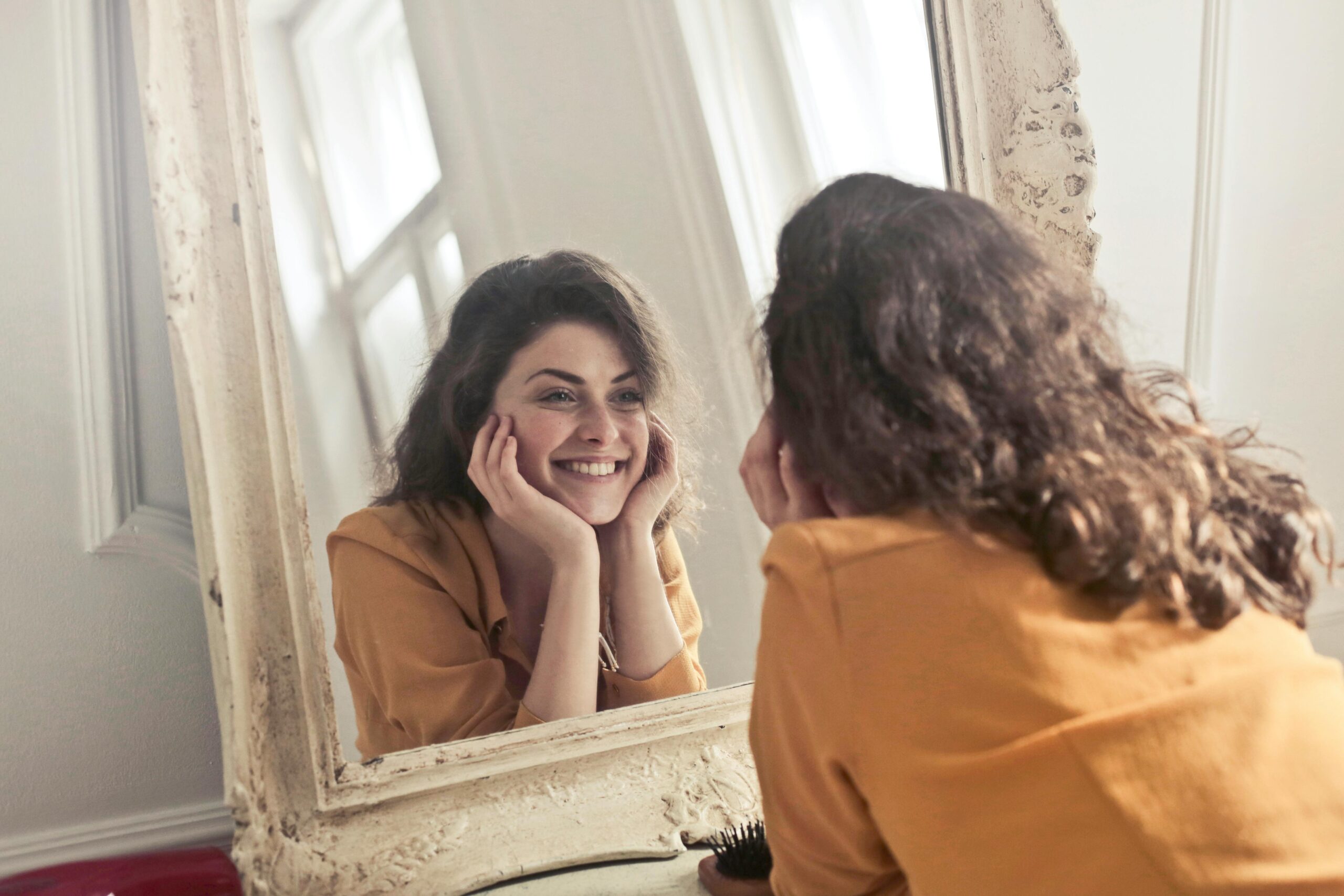 A woman with curly hair smiles authentically while looking at herself in a large, ornate mirror. She is wearing a mustard-colored top and has her hands resting on her cheeks.