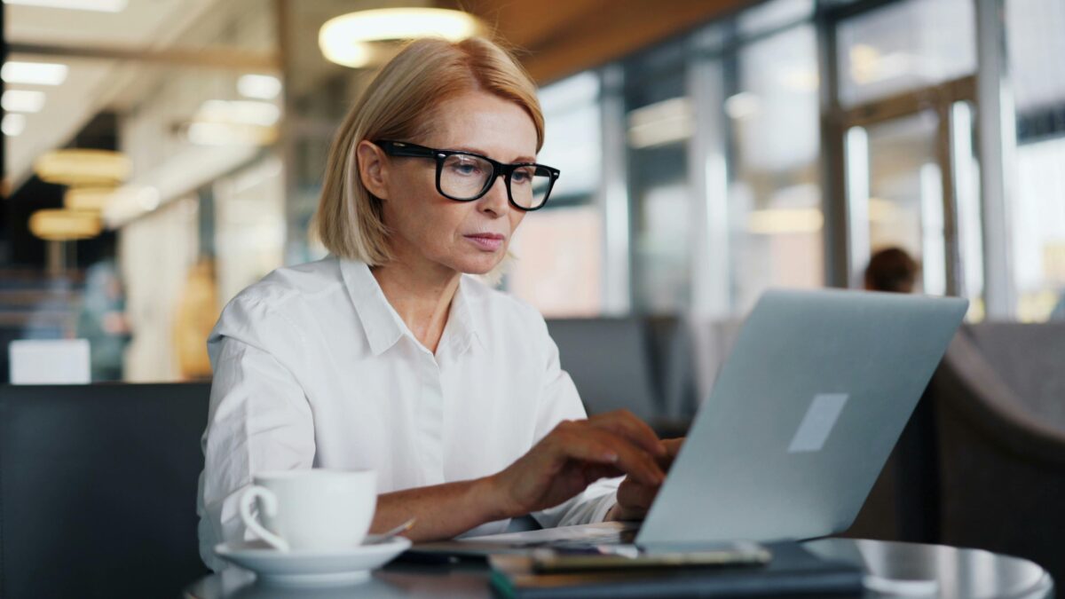 A person with short blonde hair and glasses is sitting at a table in a modern setting, working on a laptop. A cup of coffee and papers are on the table, as they follow their guide to redefining creativity in thriving midlife.