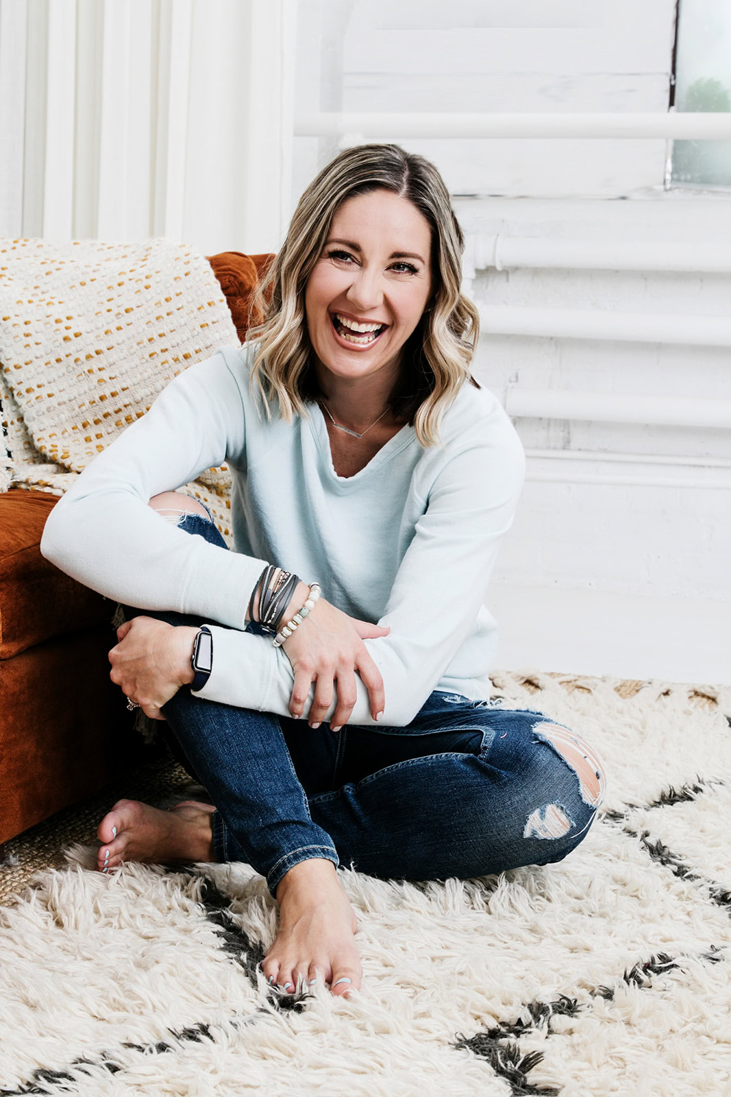 Shannon Talbot with blonde hair, wearing a light blue sweater and ripped jeans, is sitting barefoot on a plush rug, smiling contentedly. She leans against an orange couch in a bright room with white walls and windows, redefining success in her own way.