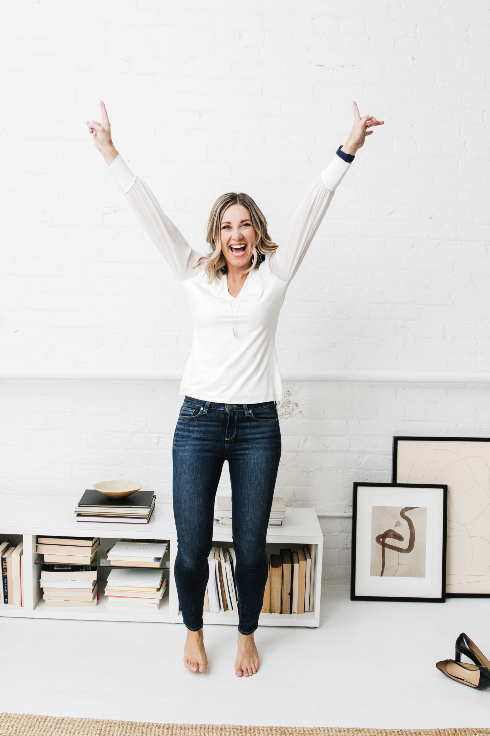 Shannon Talbot stands in the room with arms raised, smiling. The space, featuring white brick walls, bookshelves, and framed art on the floor, exudes an atmosphere of personal growth.