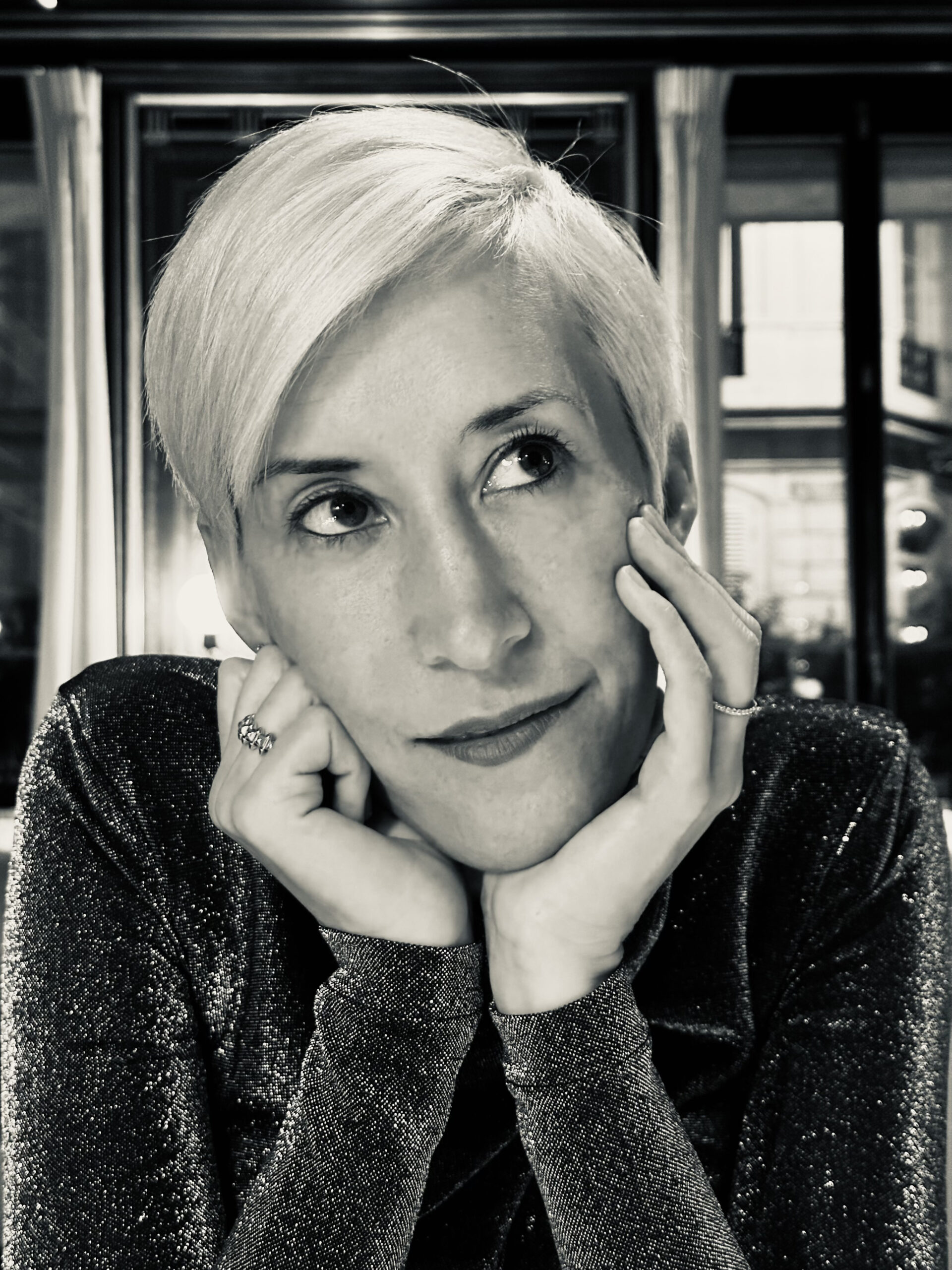 Jennifer K Hill, with short hair, rests her face in her hands, looking thoughtful. She is wearing a shimmery top. The background includes a window with night city lights.