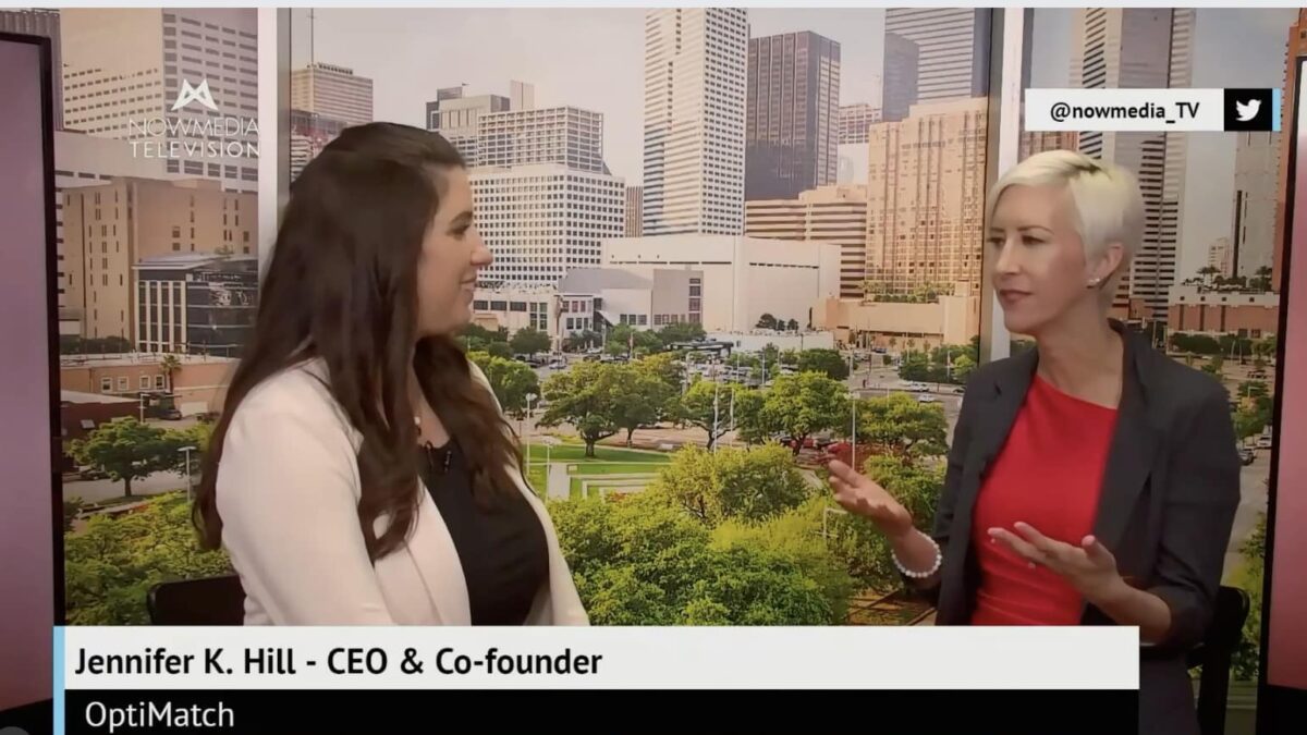 Two women, embodying resilience and flourishing in midlife, are seated in front of a cityscape background, engaged in conversation. One woman has long brown hair and the other has short blonde hair. Both are dressed in business attire.