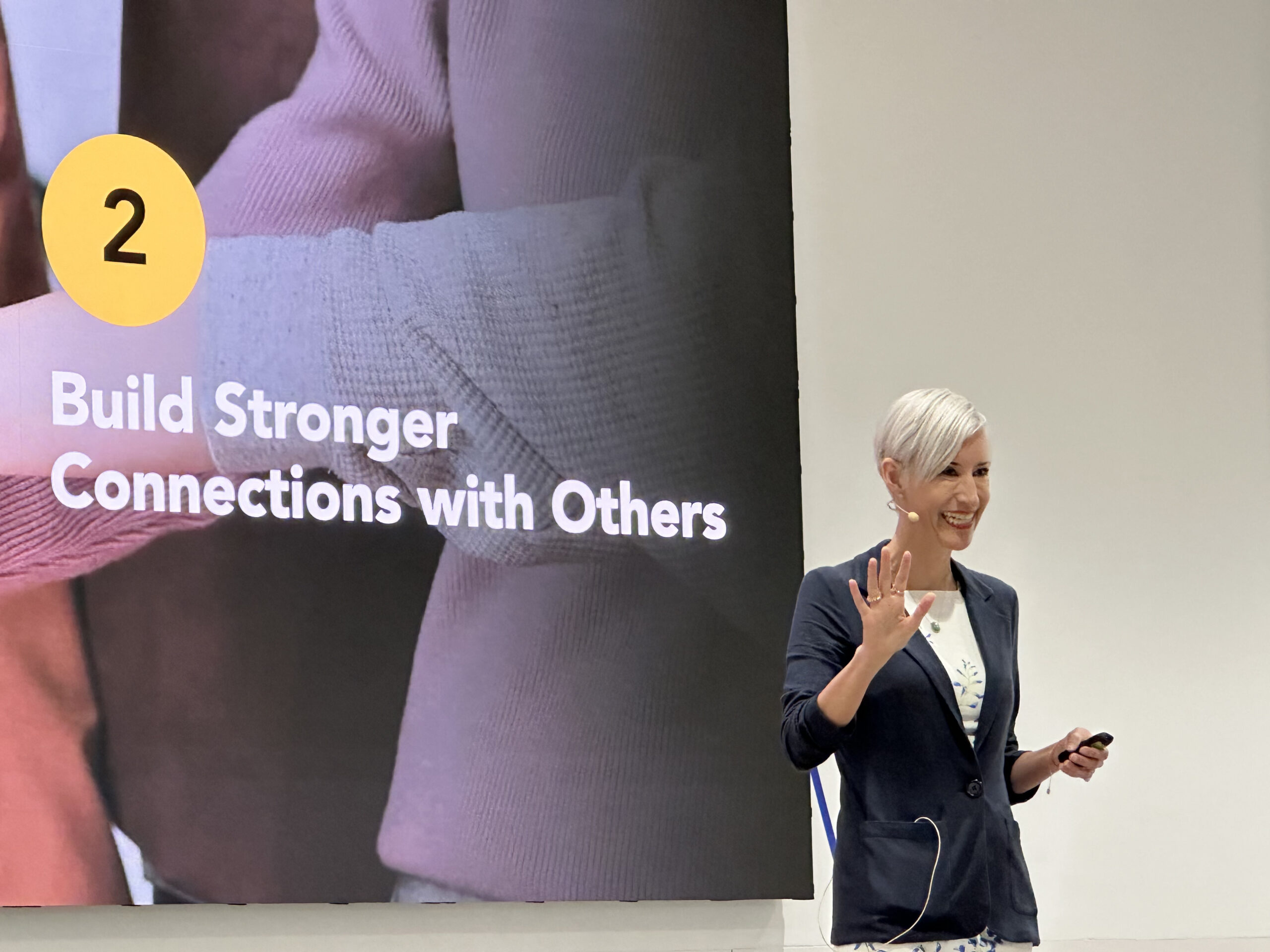 Jennifer K Hill with short hair and a dark cardigan is speaking in front of a screen displaying "2 Build Stronger Connections with Others" during a presentation on the Power-of-Innovation-in-Midlife.