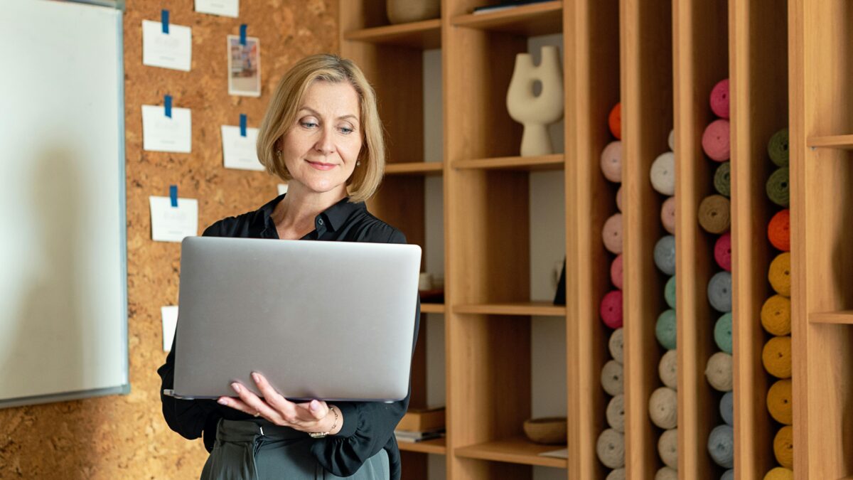 A woman stands in a craft studio, rediscovering and redefining her passions while holding a laptop and smiling. She is in front of a shelf filled with colorful yarn and a corkboard with papers clipped to it.