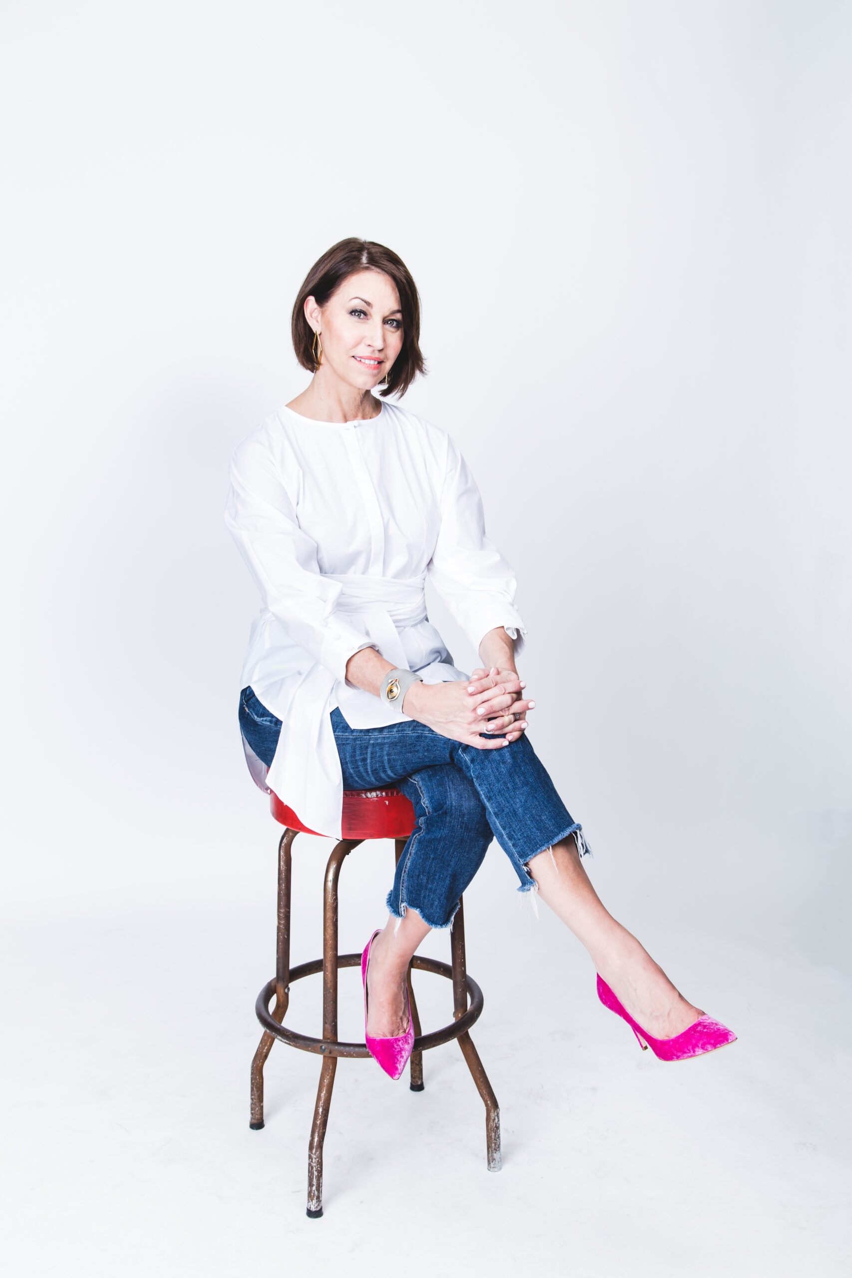 Alison Bruhn sits on a stool, dressed in a white blouse and jeans. She pairs her outfit with pink shoes and has short dark hair, all set against a white background.
