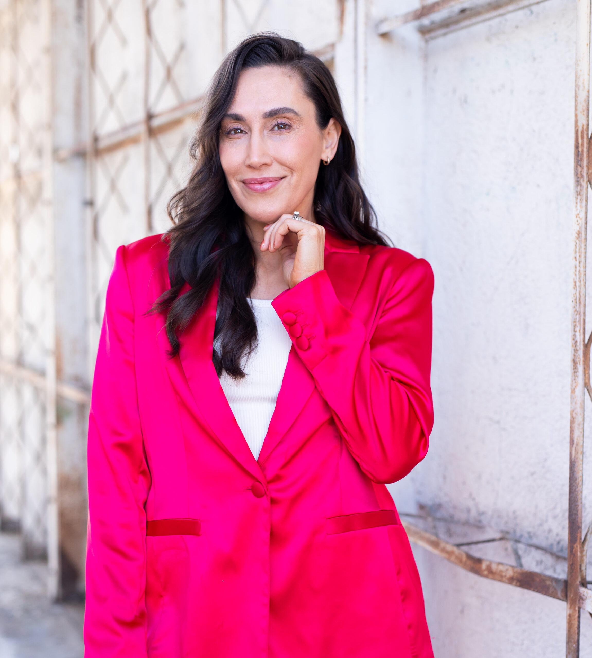 Dr. Anna Marie Frank with long dark hair wearing a vibrant pink blazer and white top stands against a light-colored, industrial-style background, smiling while gently touching their chin, embodying the spirit of empowering personal health choices.