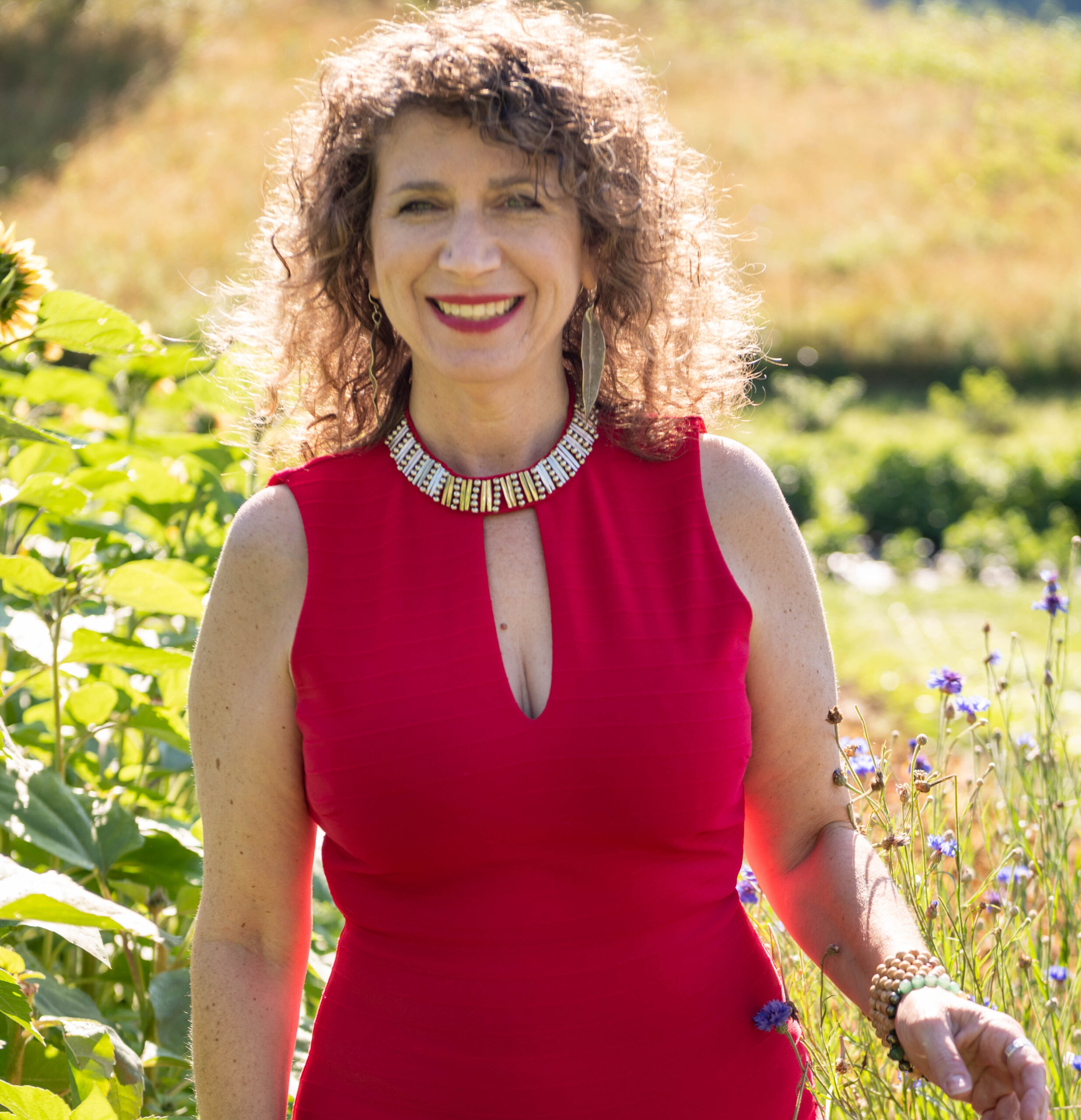 Junie Moon with curly hair standing outdoors in a garden, wearing a red dress and a statement necklace, smiling at the camera Betting on Love.