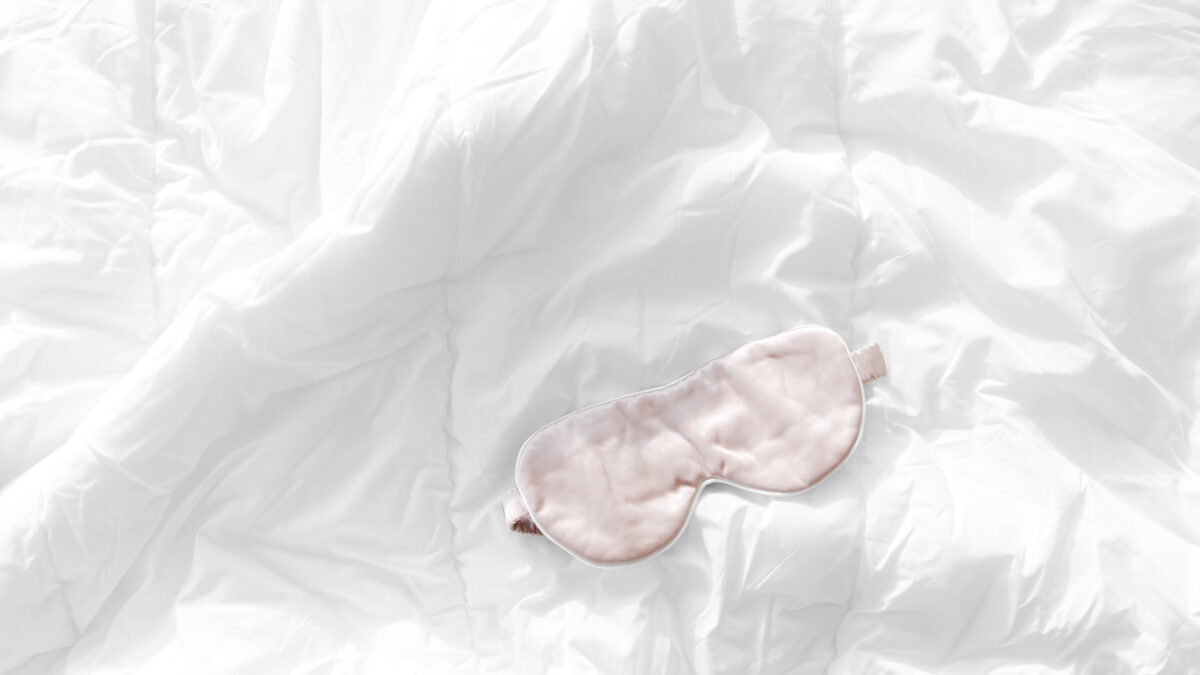 A pink eye mask, symbolizing the crucial role of sleep, lies on a white crumpled bed sheet.