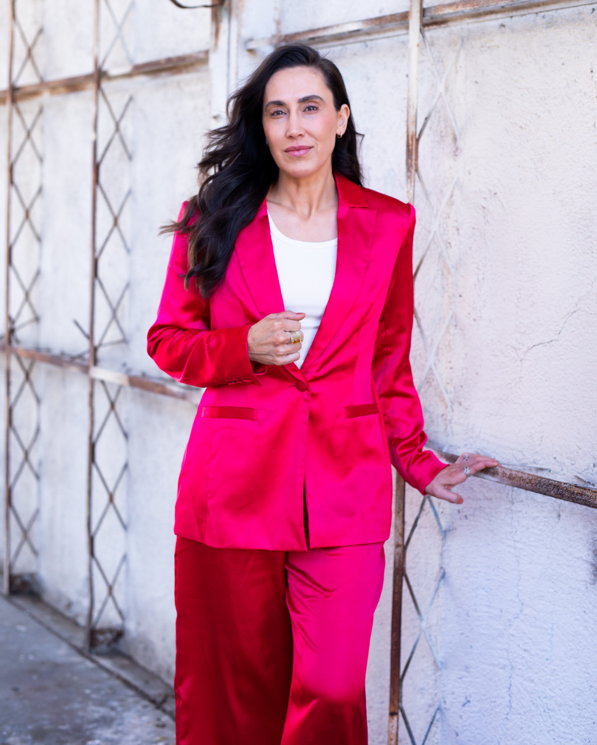 Dr. Anna Marie Frank wearing a bright pink satin suit stands against a white, crisscross-patterned wall. She has long, dark hair and holds onto the wall with one hand, embodying strength and resilience while combatting mental health challenges.