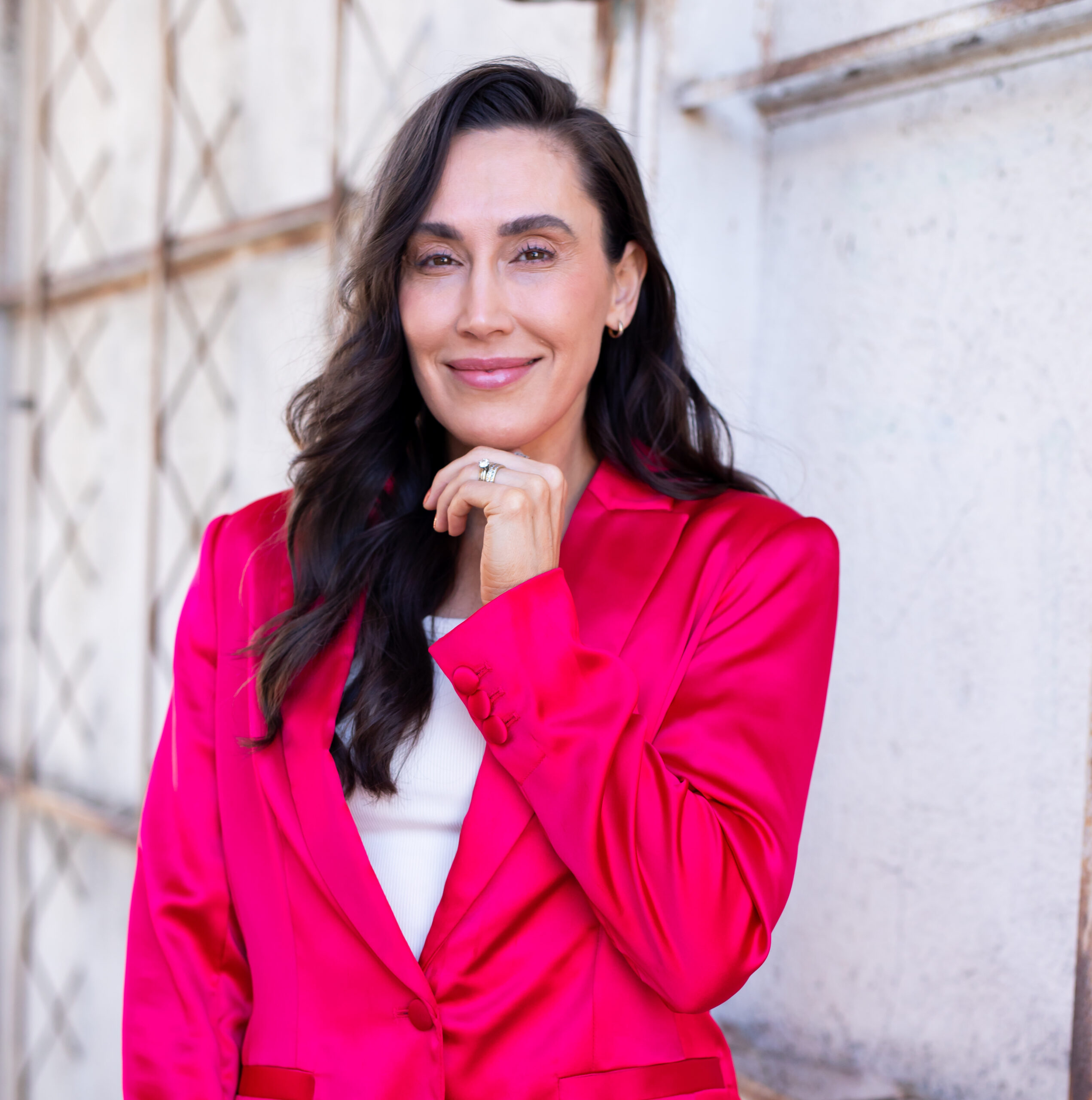 Dr. Anna Marie Frank with long brown hair is wearing a bright pink blazer and smiling at the camera. She has one hand resting near her chin and is standing against a light-colored wall with a crisscross pattern, radiating confidence akin to someone who advocates for natural remedies.