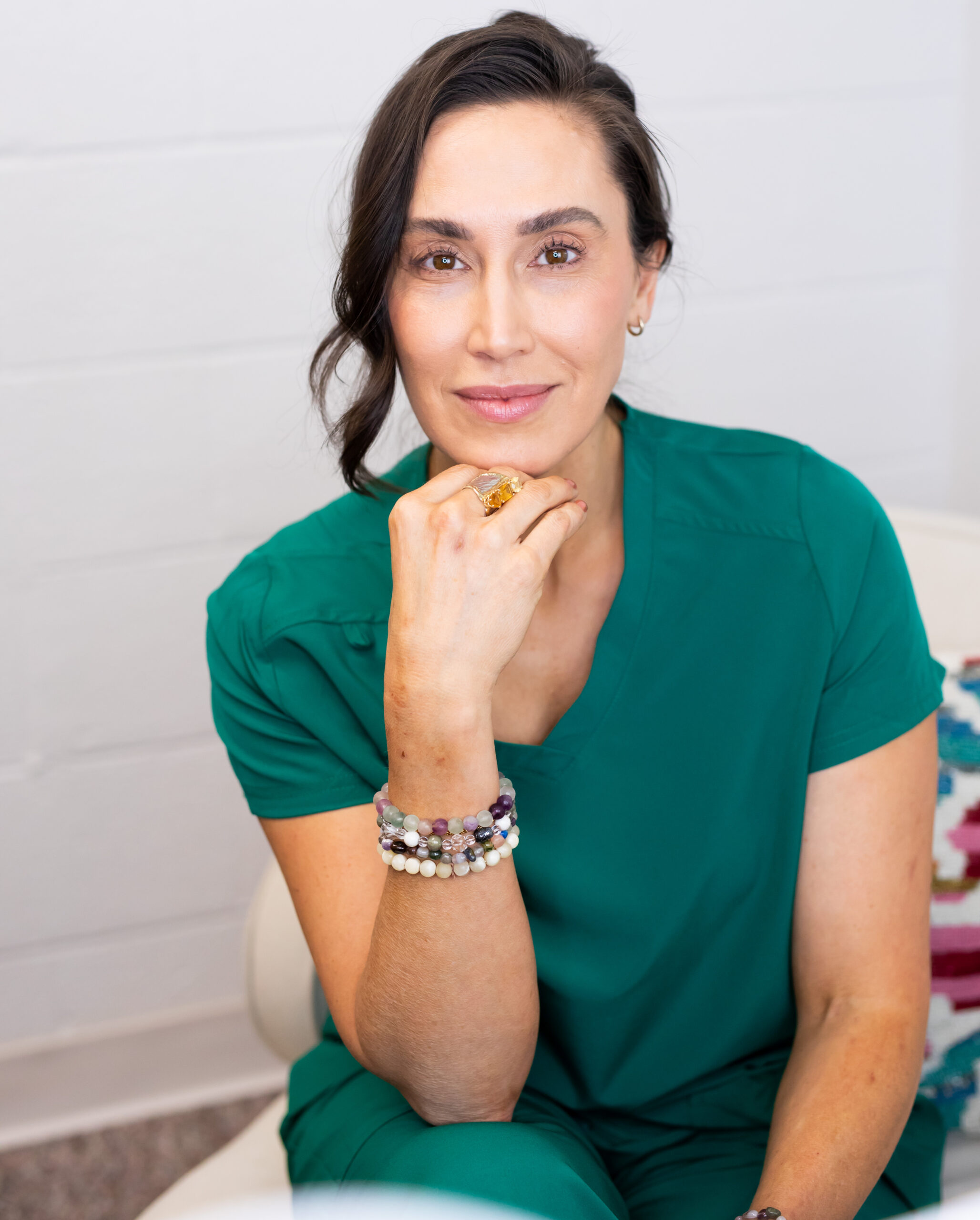 Dr. Anna Marie Frank, wearing a green top and multicolored beaded bracelets, sits with her hand thoughtfully under her chin against a white background.