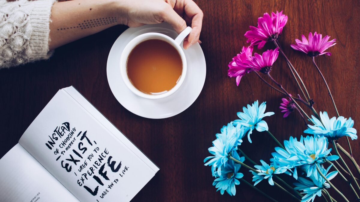 A hand holding a cup of tea near an open book with a motivational quote and a bouquet of pink and blue flowers on a wooden table, emphasizing the impact of a supportive community in midlife.