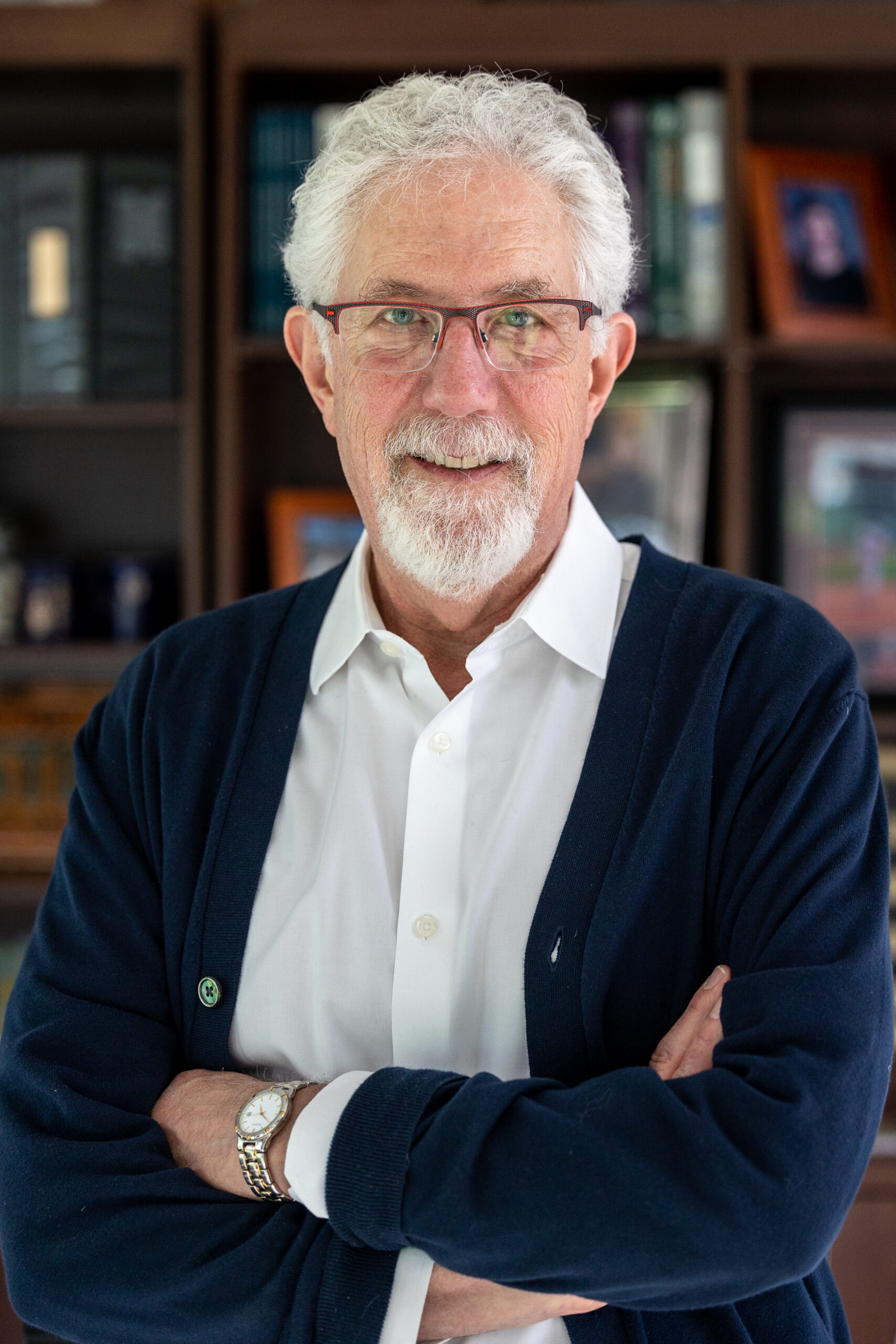 Dr. Mitchell Clionsky with white hair and glasses stands with his arms crossed, wearing a white shirt and dark cardigan, in front of a bookcase filled with books and framed photos. Meet our upcoming guest, Dr. Mitchell Clionsky.